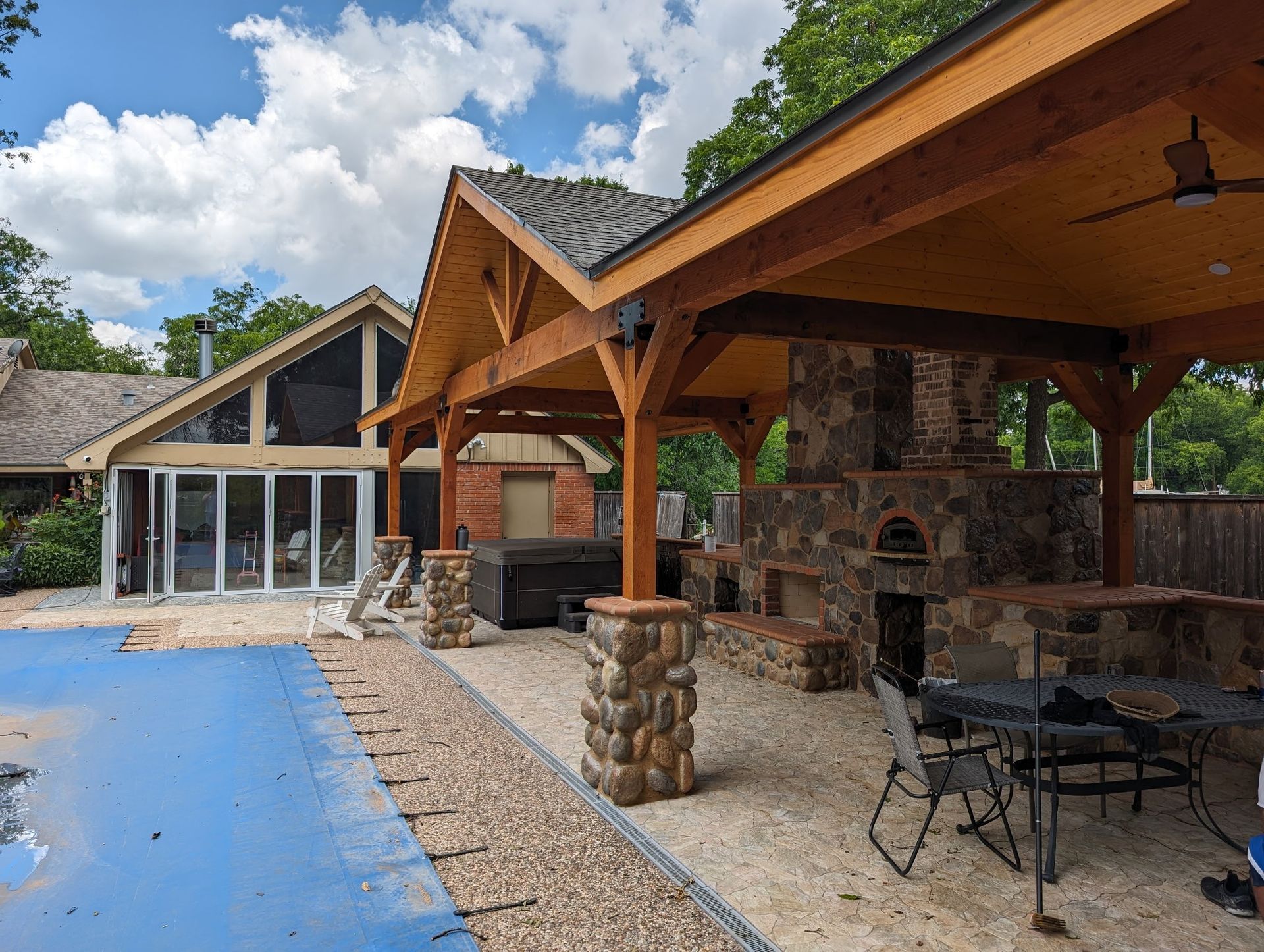 Backyard with a stone fireplace, outdoor kitchen under a wooden canopy, pool, and house.