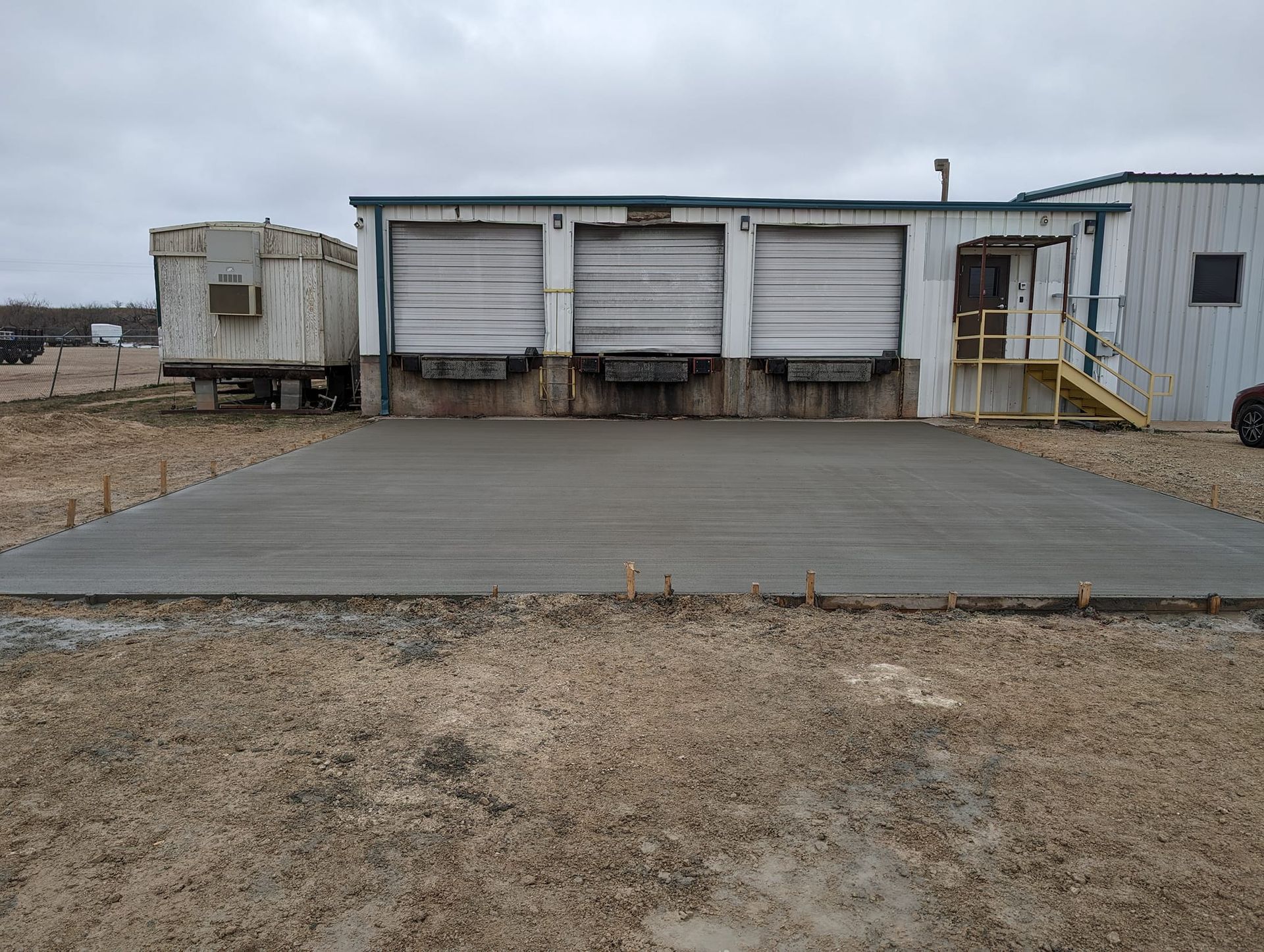 Concrete loading dock with three garage doors, a trailer to the left, and a ramp, in a field.