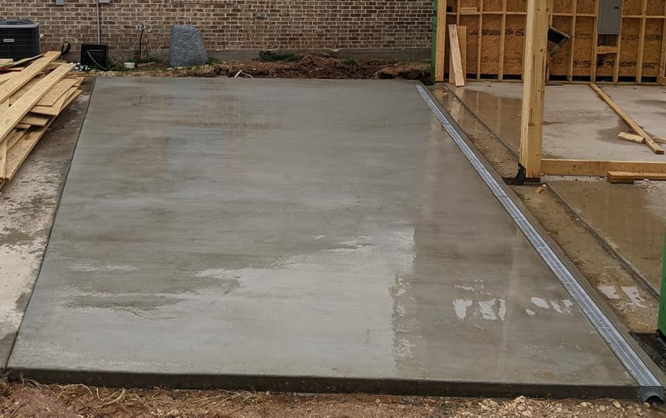Concrete slab with a drain, next to a wood frame building under construction, outdoors.