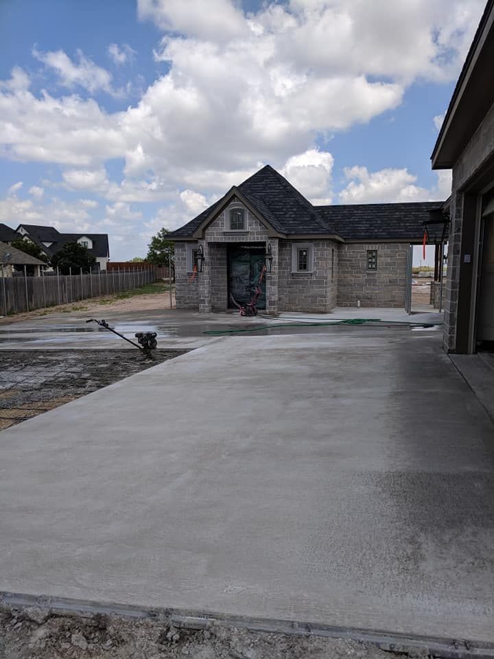 New concrete driveway leading to a light brick house with a dark roof, under a cloudy blue sky.