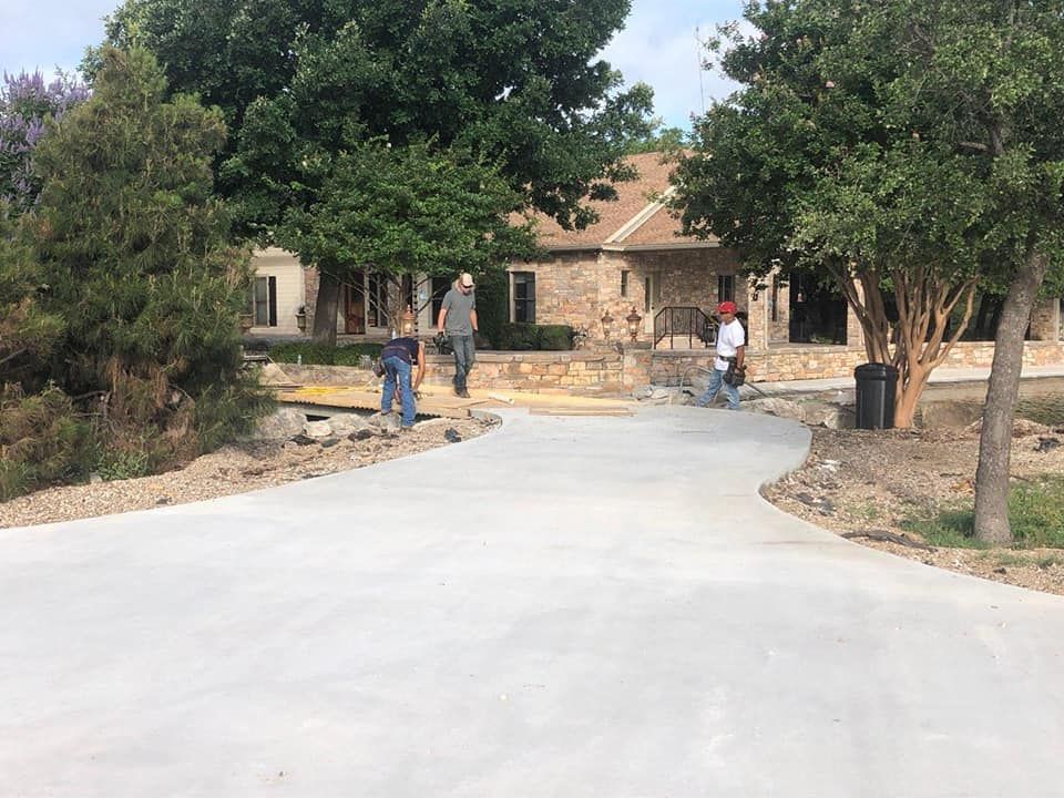 Workers near a house with a concrete driveway, surrounded by trees.
