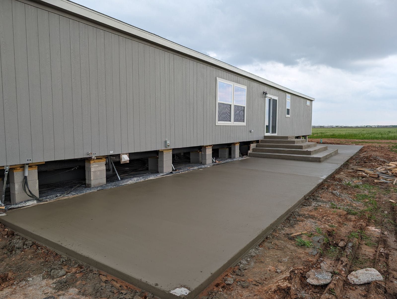 Gray mobile home with new concrete patio and steps. Cloudy sky and dirt ground.