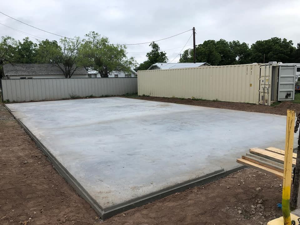 Concrete slab foundation in dirt yard next to fence and container, cloudy sky.