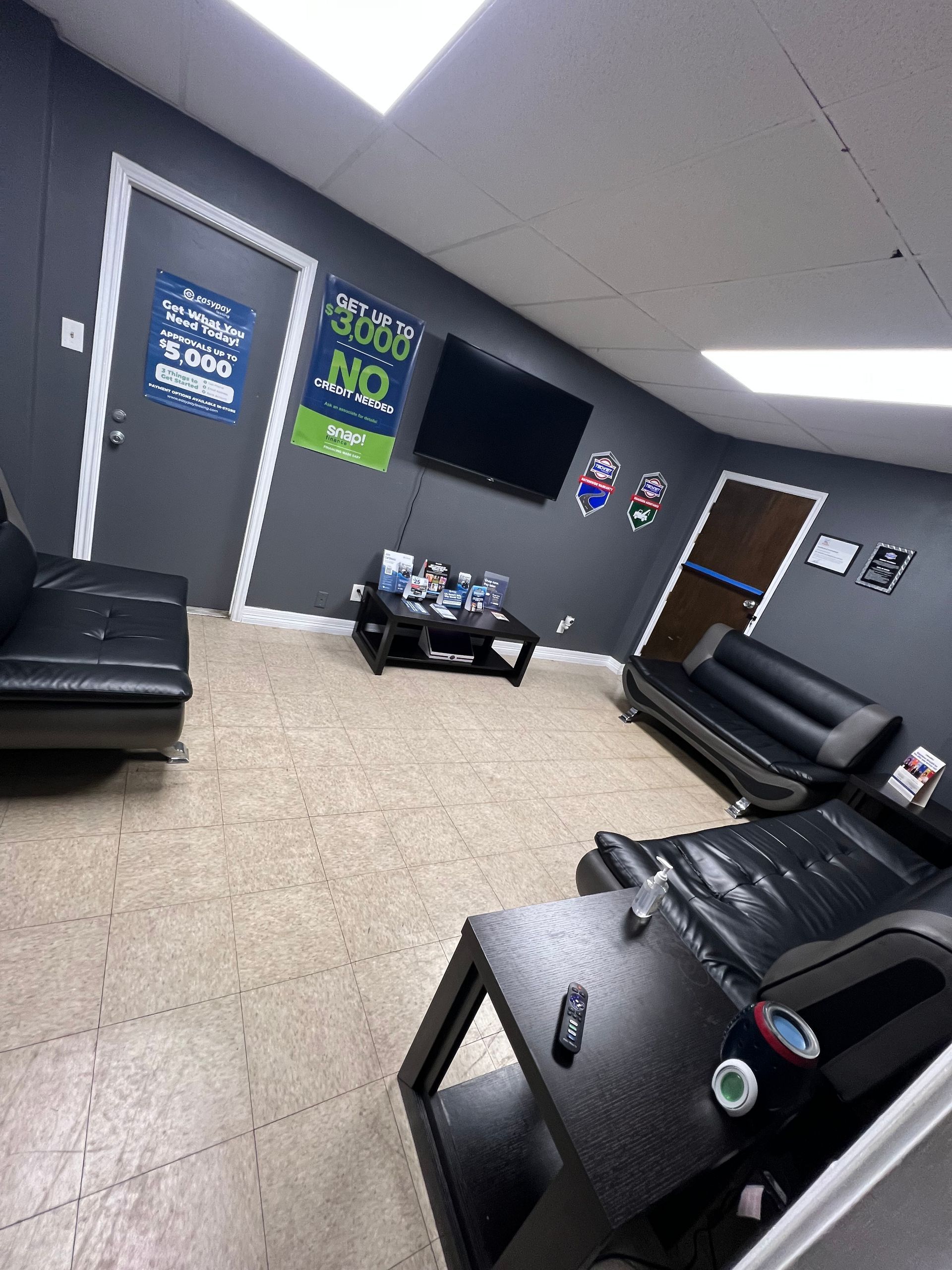 Waiting room with black leather couches, a coffee table, and a TV on a gray wall. | J & J Elite Auto Repair