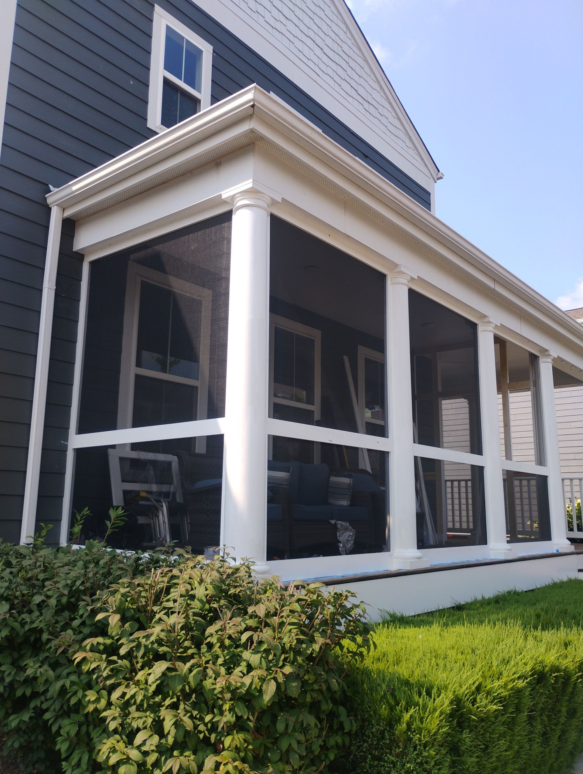 Screened-in porch with white columns, attached to a gray house, with dark screening. Sunny day, green bushes in front.