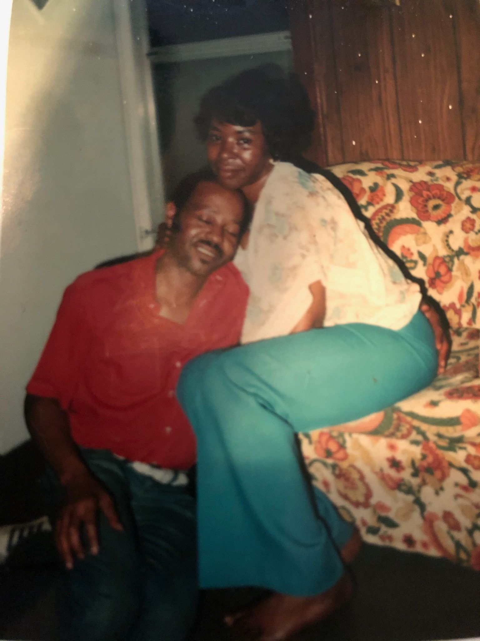 Man and woman in casual wear, seated closely on floral couch. Man in red shirt smiles, woman in teal pants smiles.