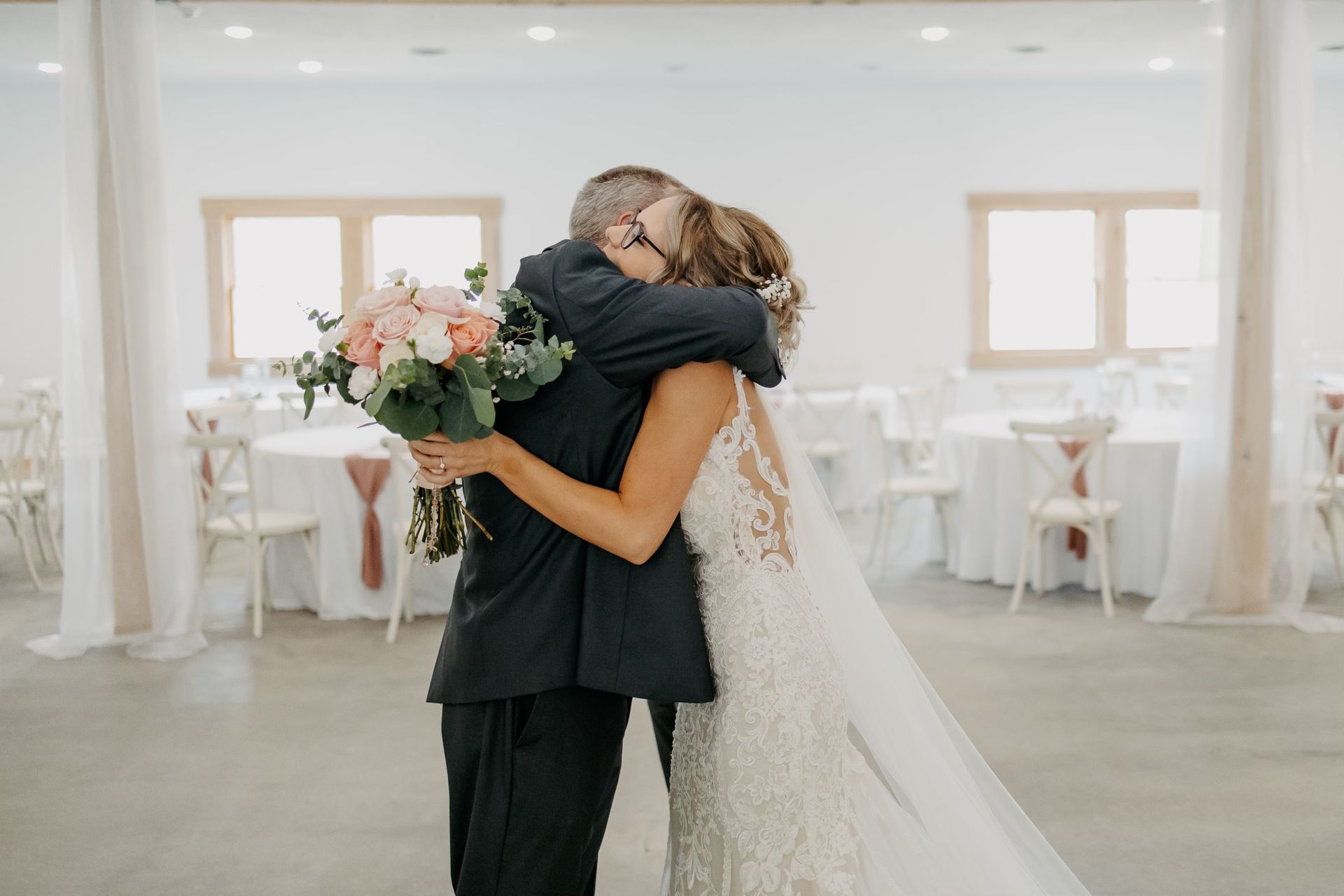 A bride and groom are hugging each other in a room . the bride is holding a bouquet of flowers.