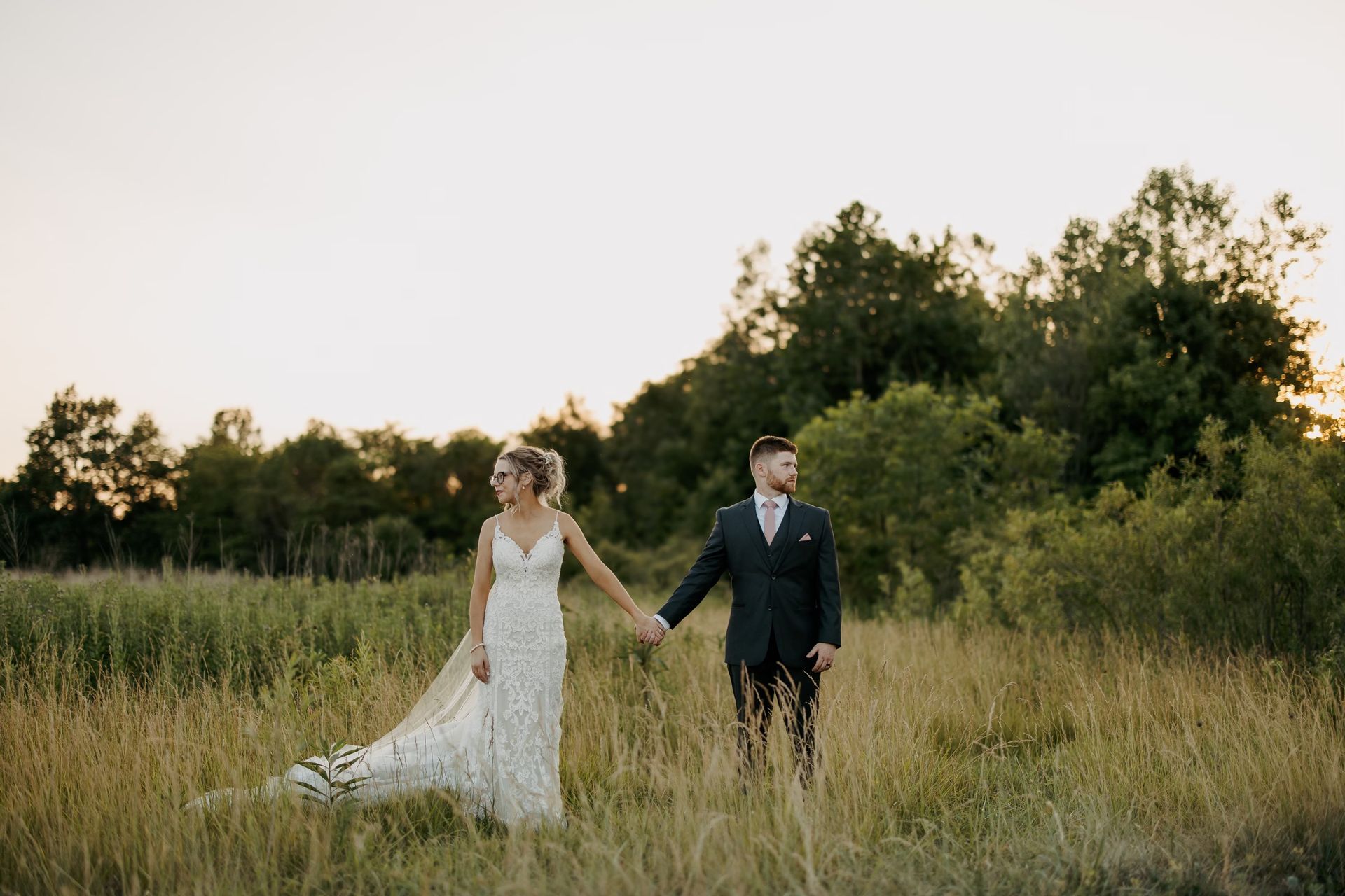 A bride and groom are walking through a field holding hands.