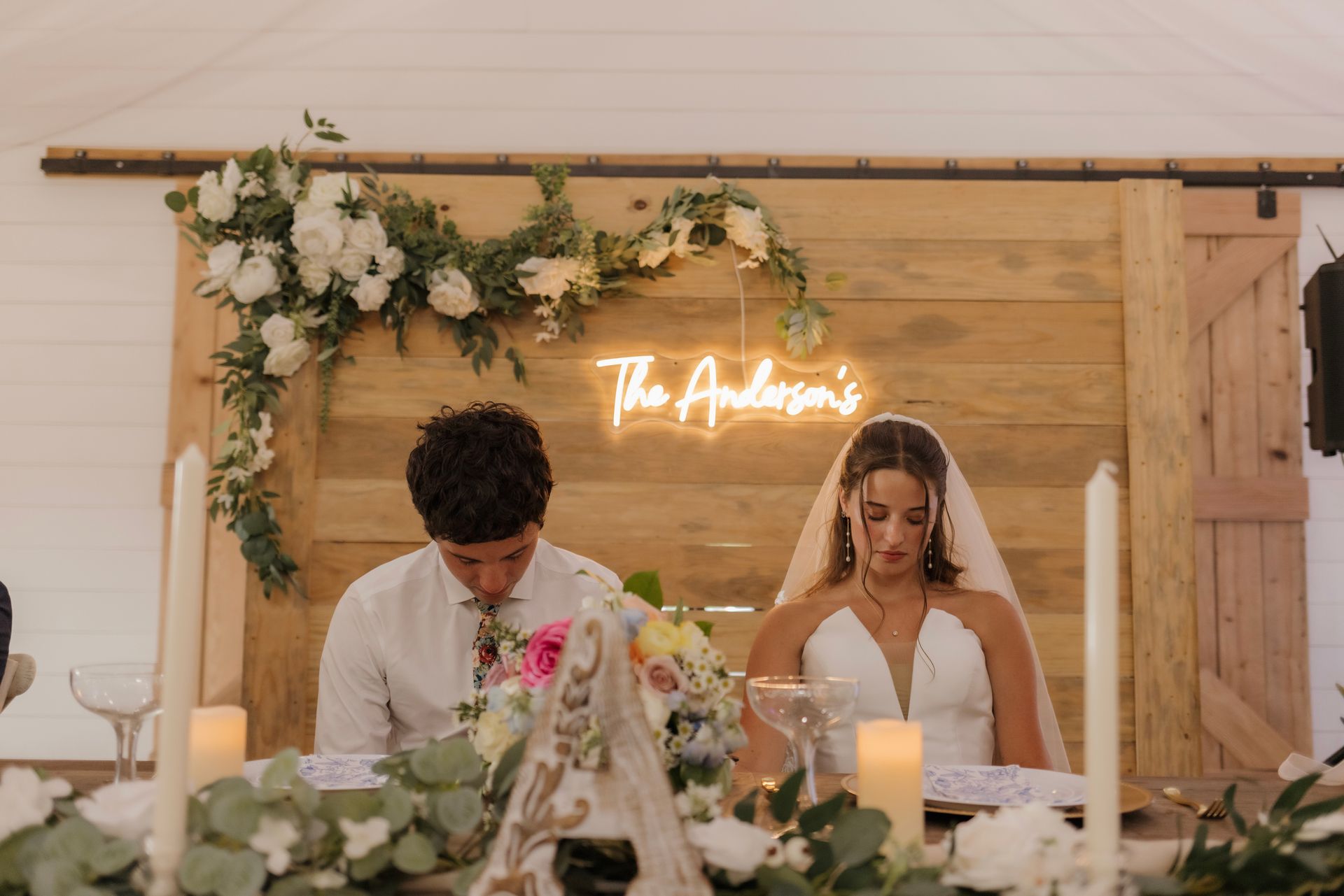 A bride and groom are sitting at a table with a neon sign that says `` the anderson 's ''.