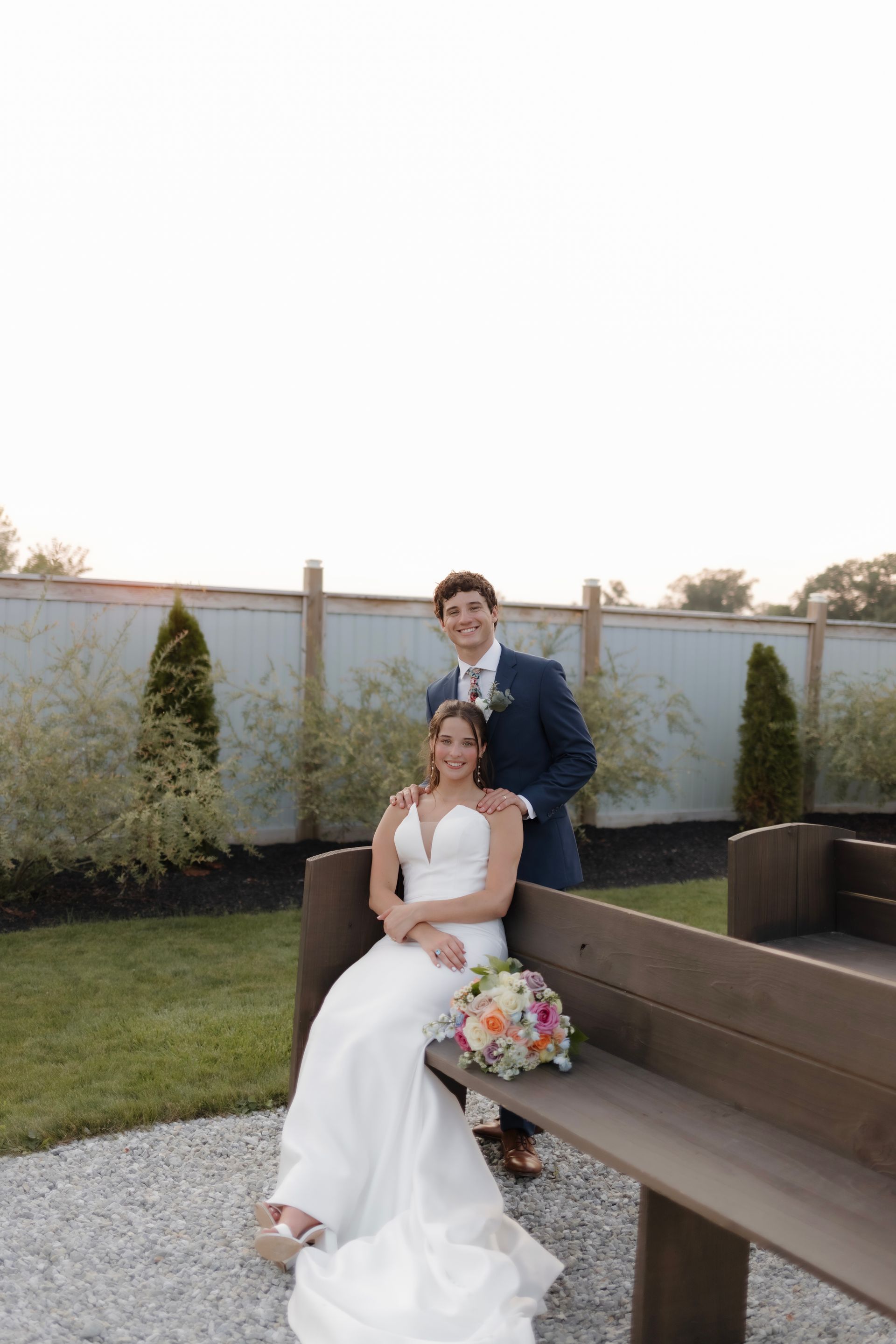 A bride and groom are posing for a picture while sitting on a wooden bench.