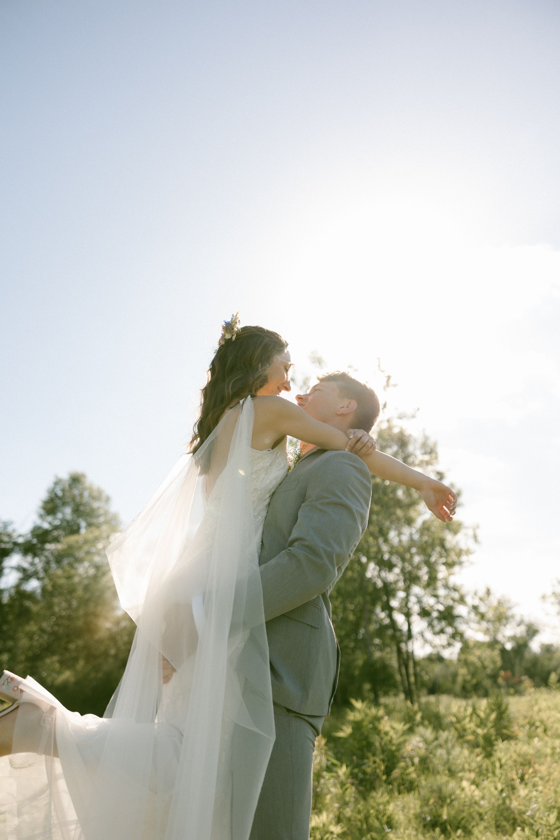 A bride and groom are kissing in a field while the groom holds the bride in his arms.
