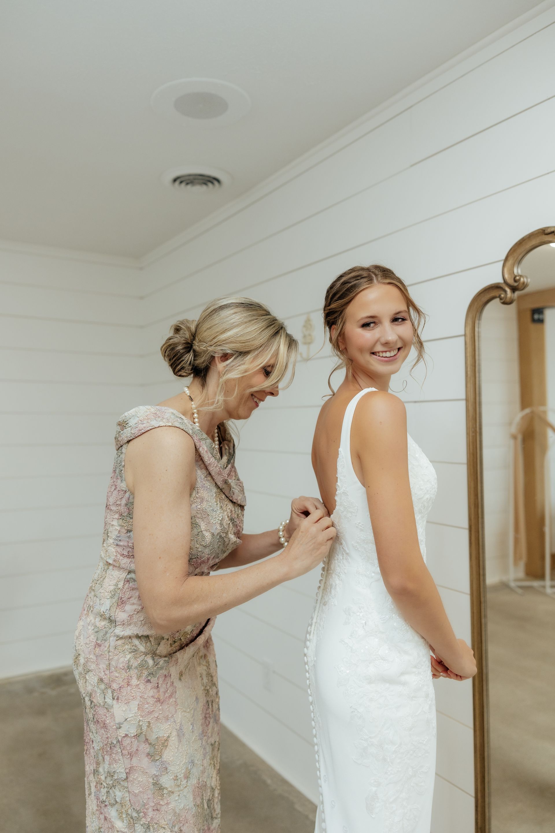 A woman is helping a bride get ready for her wedding in front of a mirror.