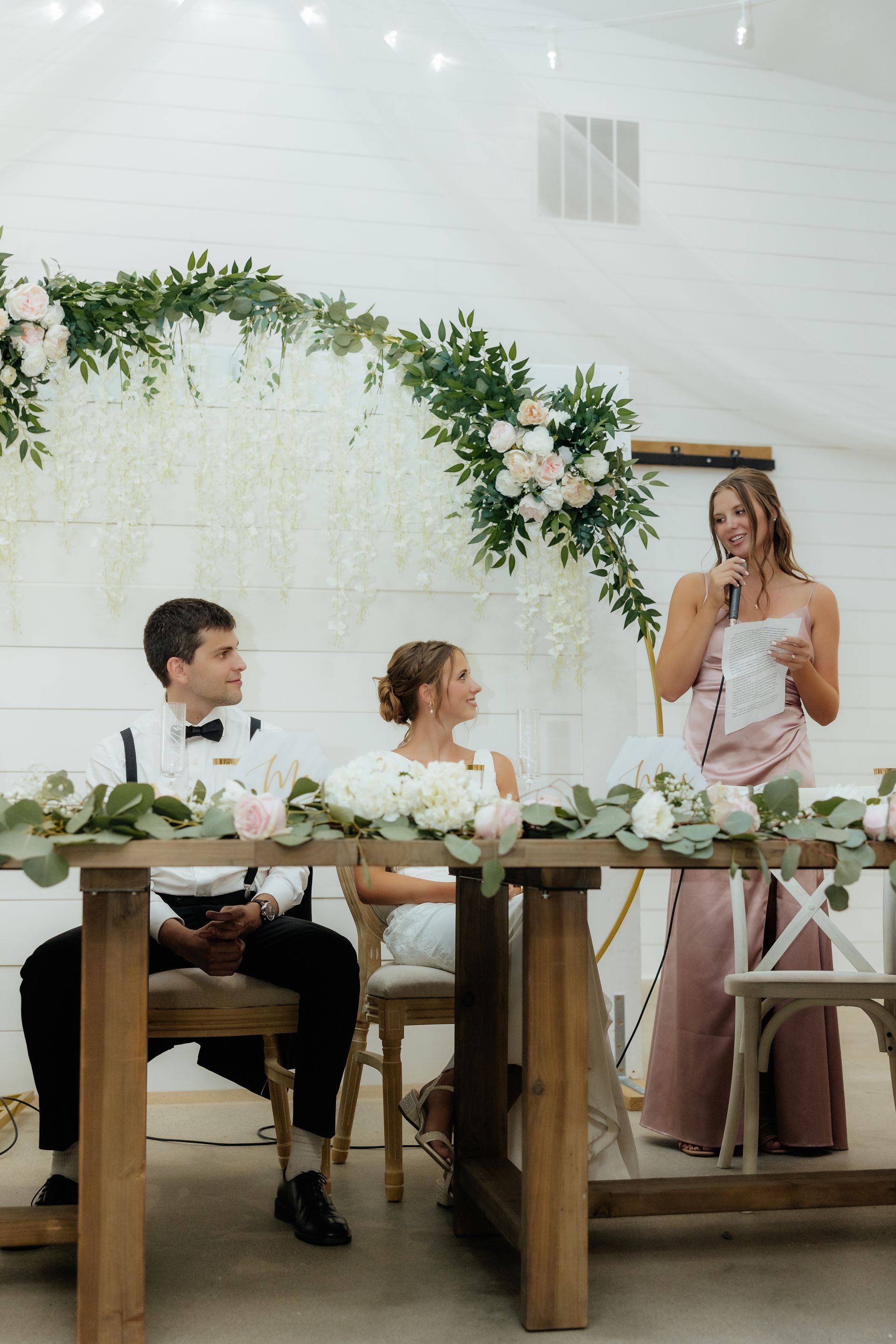A woman is giving a speech at a wedding reception while a bride and groom sit at a table.