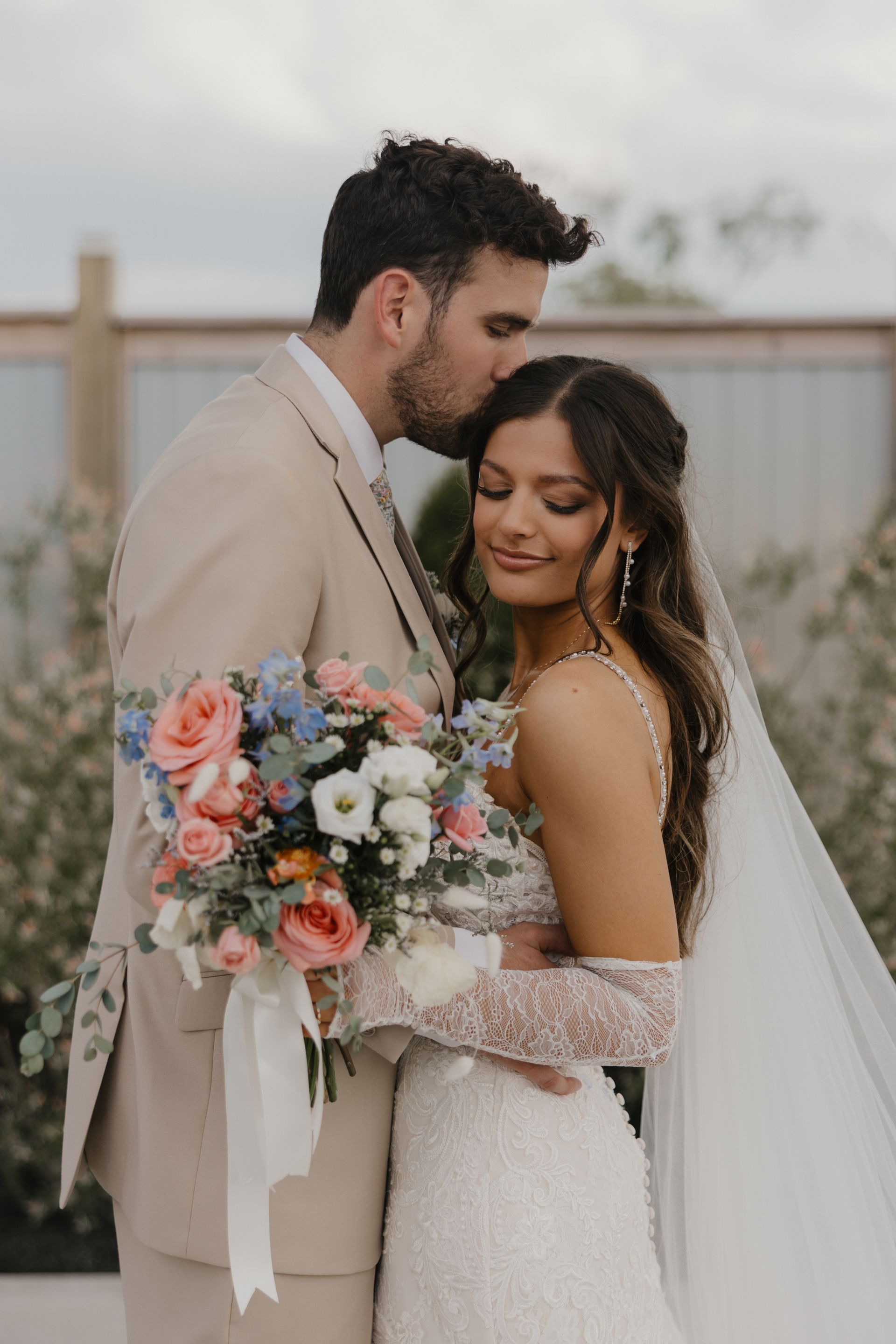 A bride and groom are posing for a picture and the groom is kissing the bride on the forehead.