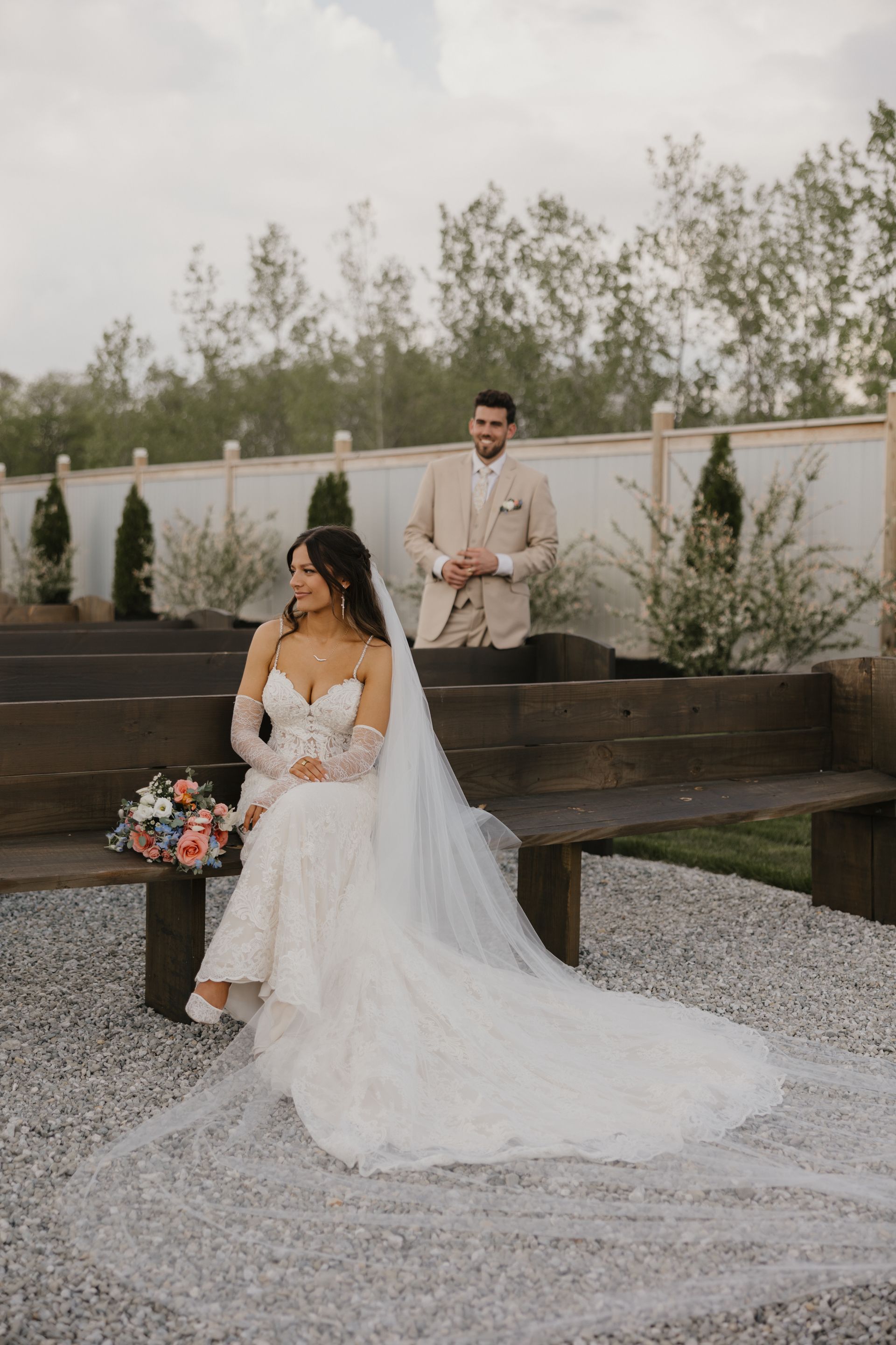 A bride and groom are posing for a picture while the bride is sitting on a bench.