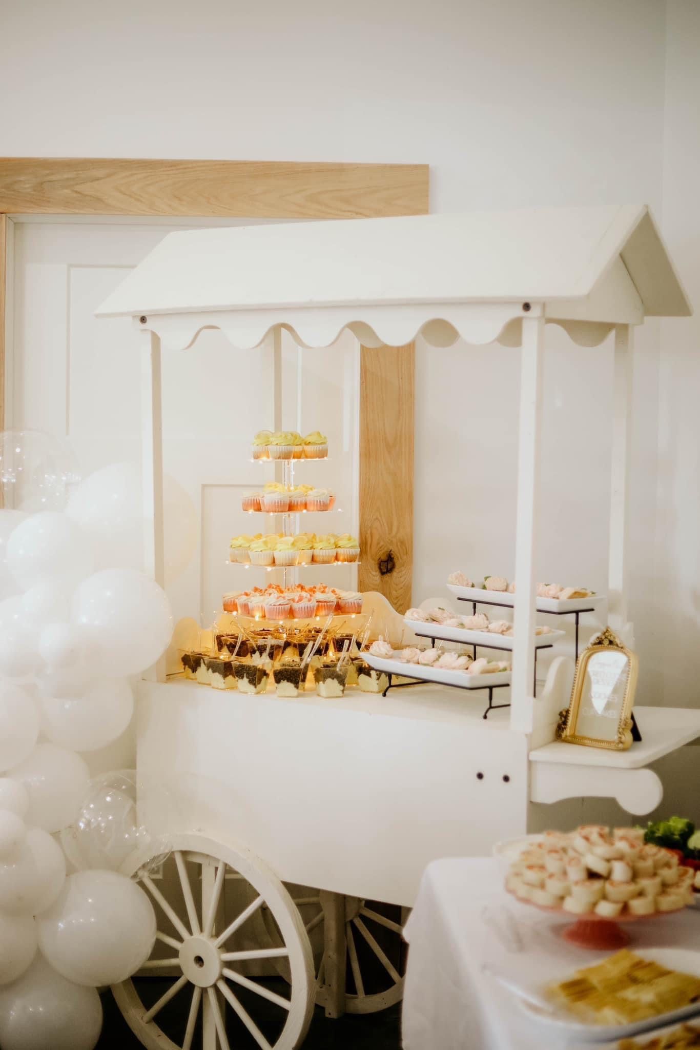 A white cart filled with cupcakes and balloons is sitting on top of a table.