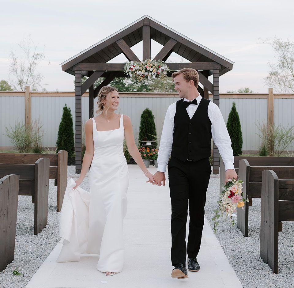 A bride and groom are walking down a white aisle holding hands