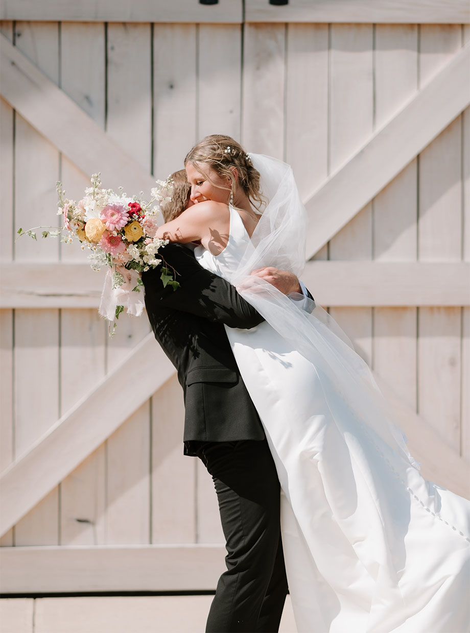 A bride and groom are hugging in front of a white barn.