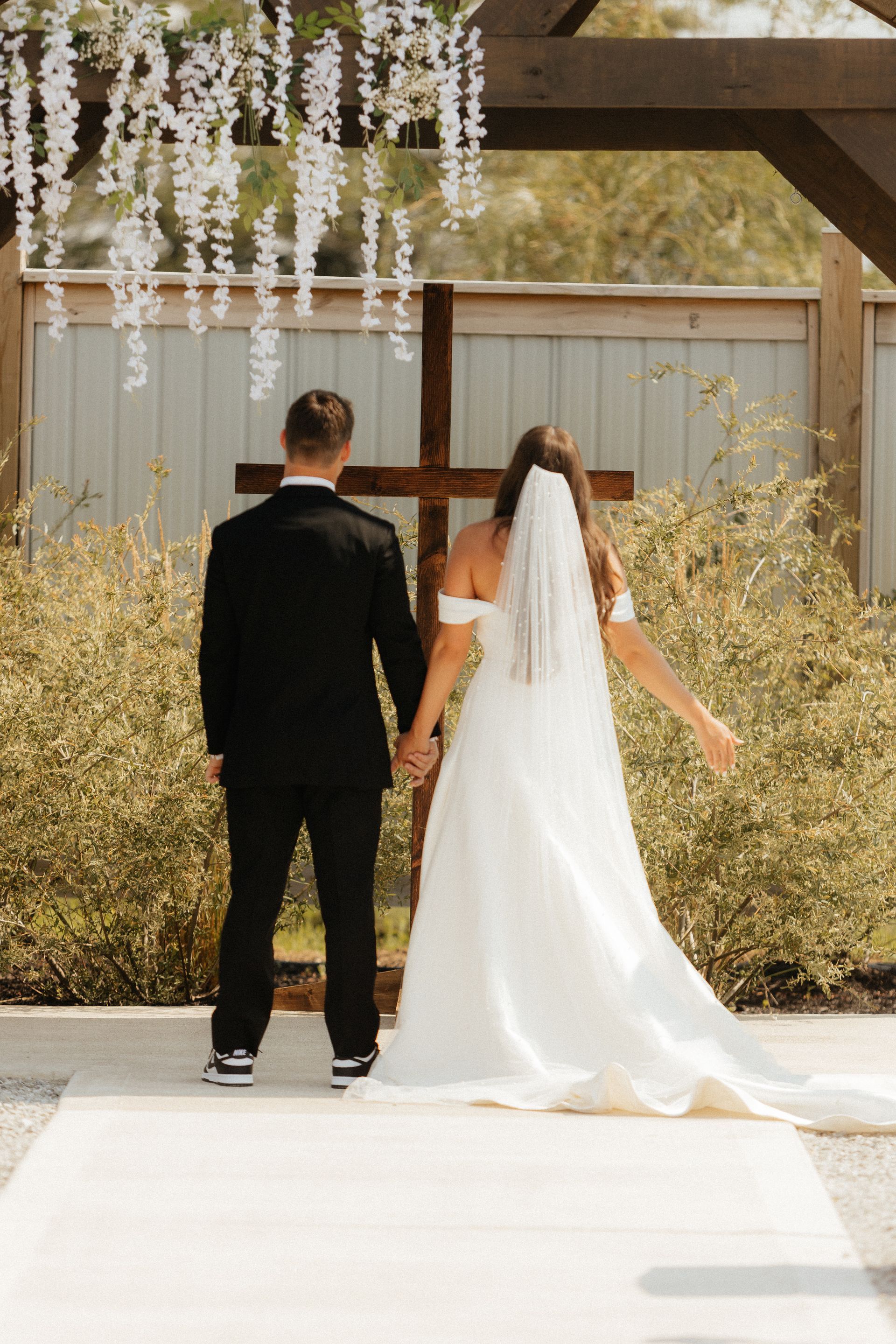 A bride and groom are standing in front of a wooden cross holding hands.