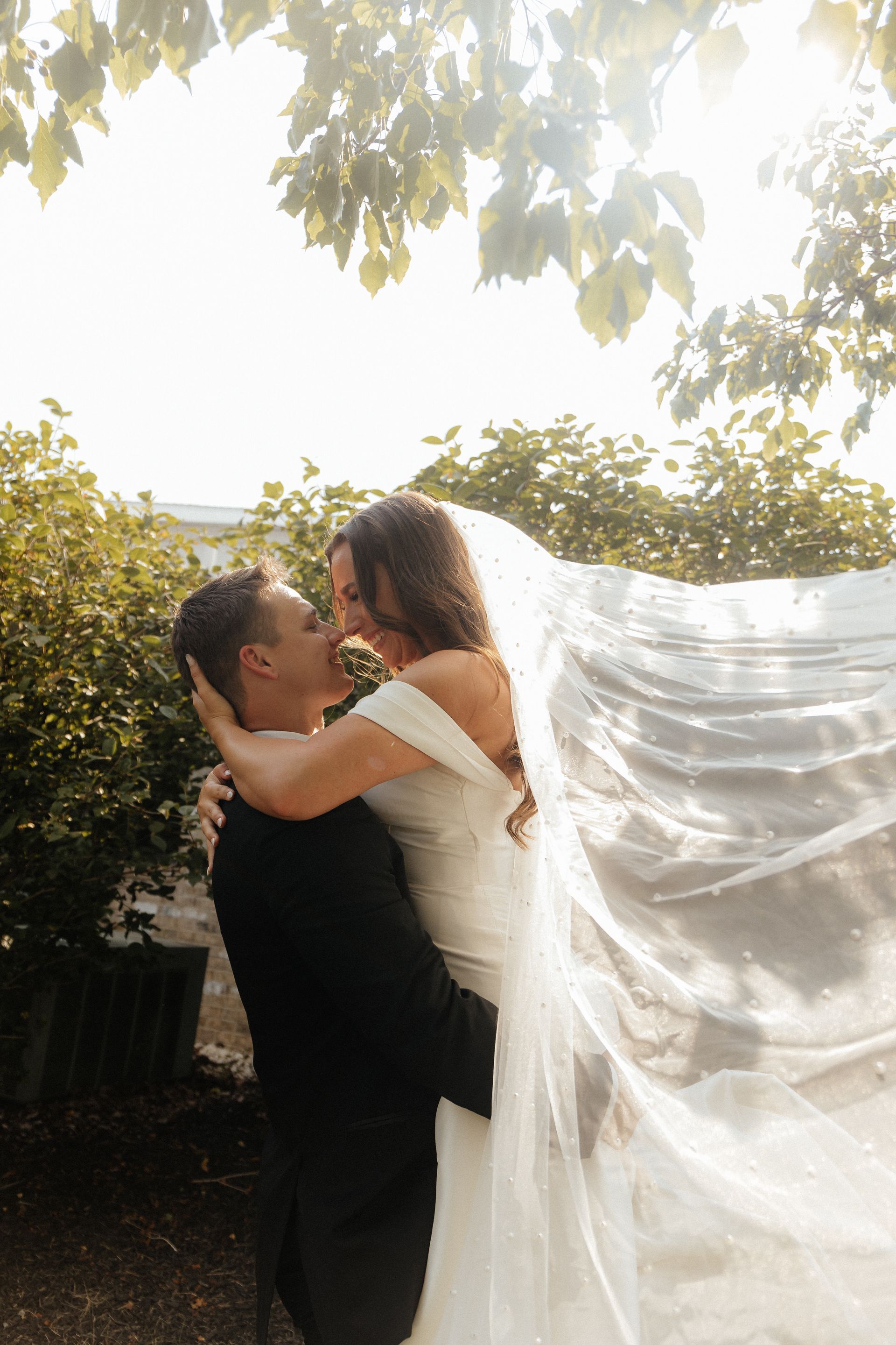 A bride and groom are kissing under a tree with a veil blowing in the wind.