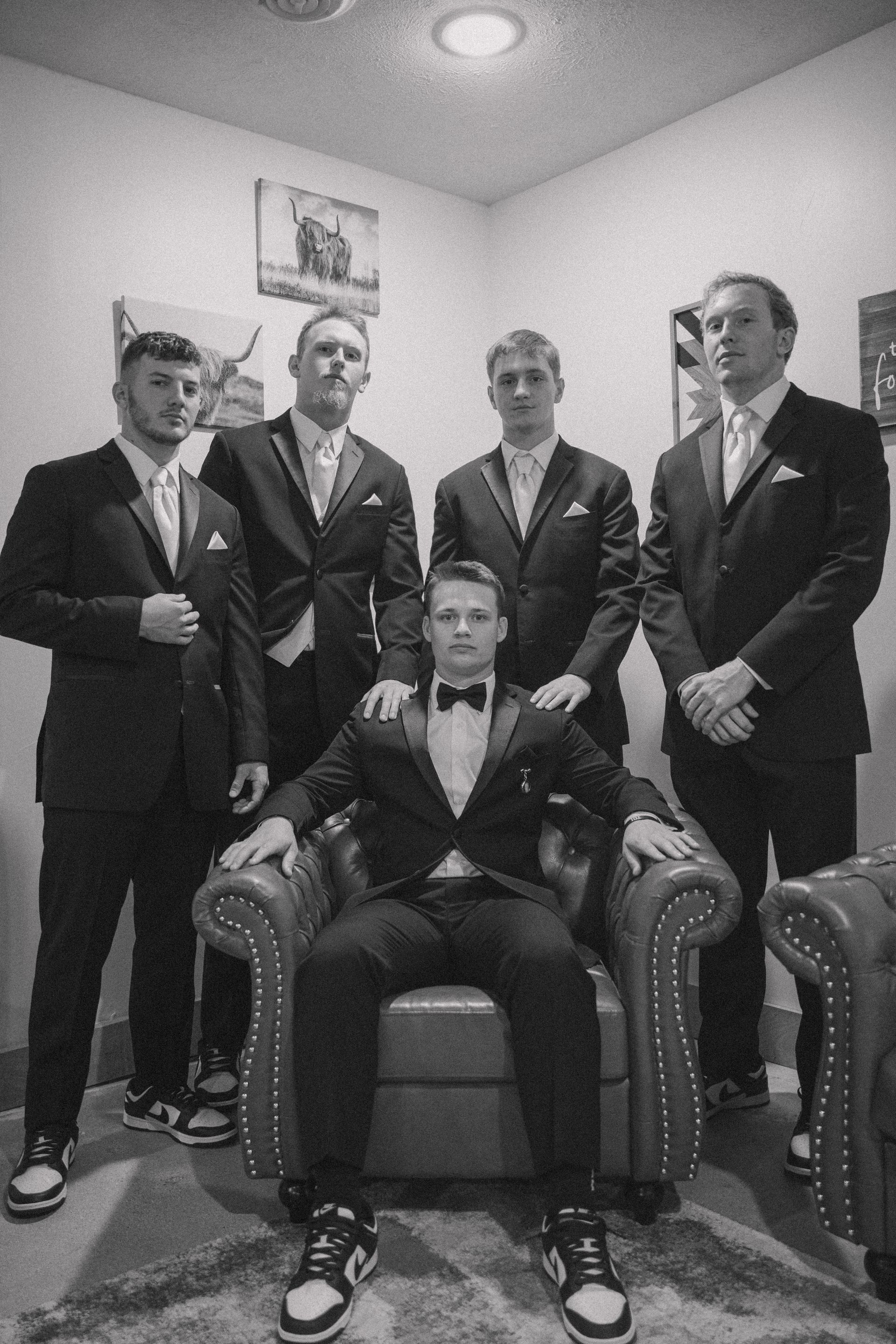 A man in a tuxedo is sitting in a chair with his groomsmen standing around him.