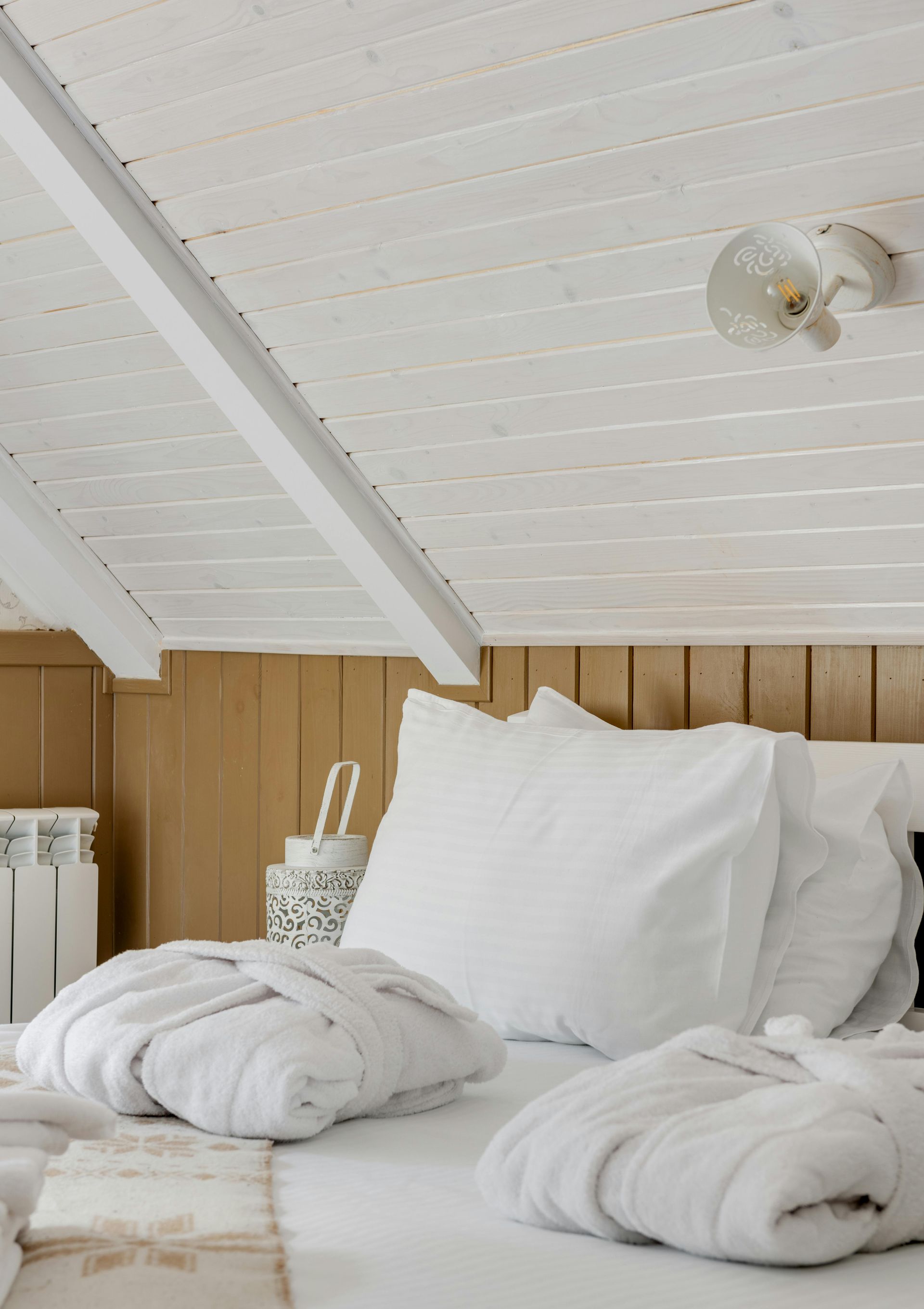 White bed with pillows and folded robes, against wood paneling and a white sloped ceiling.