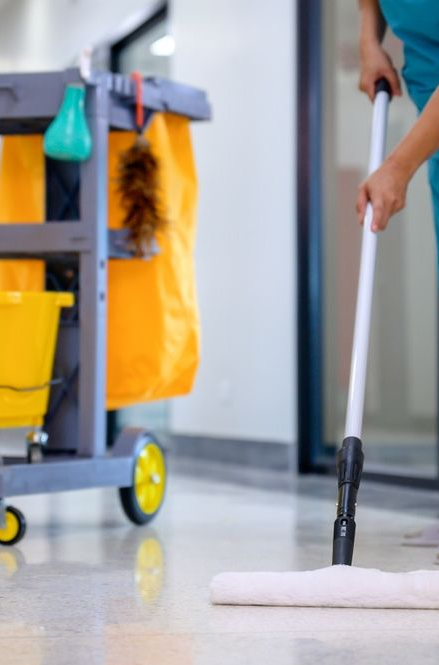 A person mopping a shiny floor with a cleaning cart beside them, which holds supplies such as a yellow bucket.