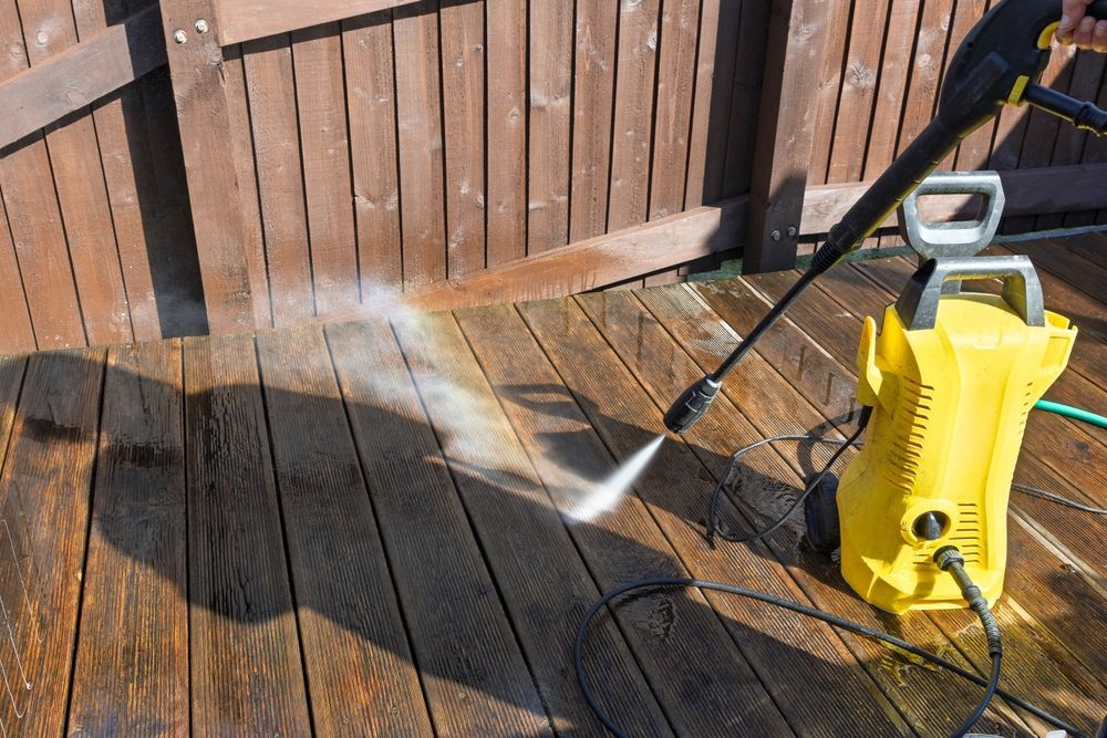 A yellow pressure washer sprays water on a wooden deck next to a fence.