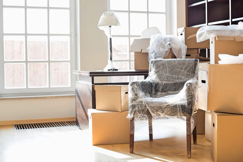 Room with moving boxes, wrapped chair, desk with lamp, and large windows.
