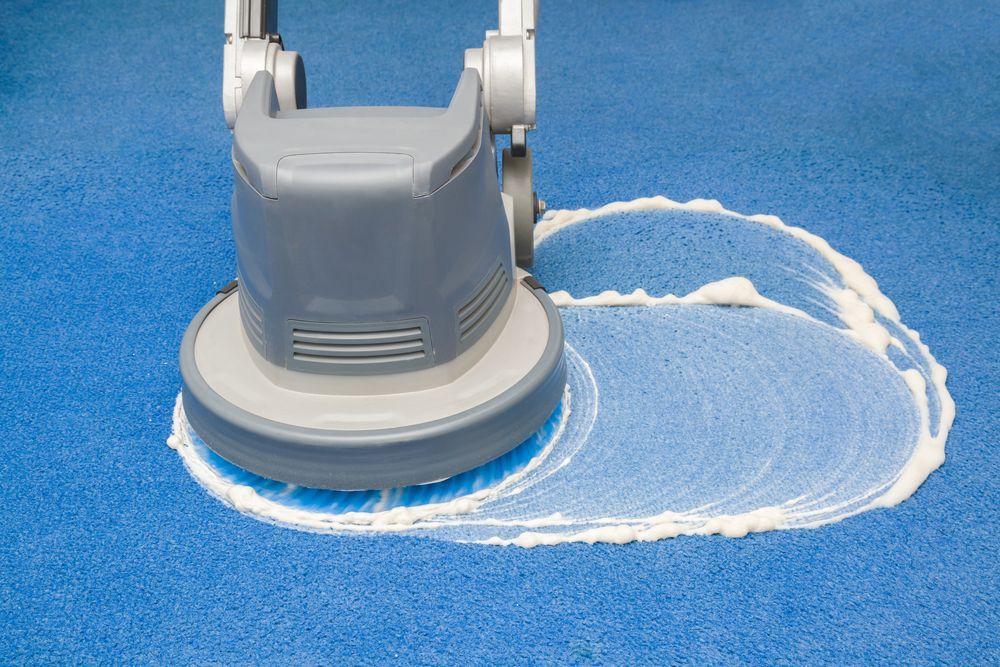 Gray floor cleaning machine scrubbing blue carpet with white foam.