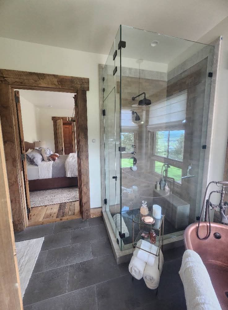 Bathroom with glass shower, copper tub, and view of a bedroom through a doorway.