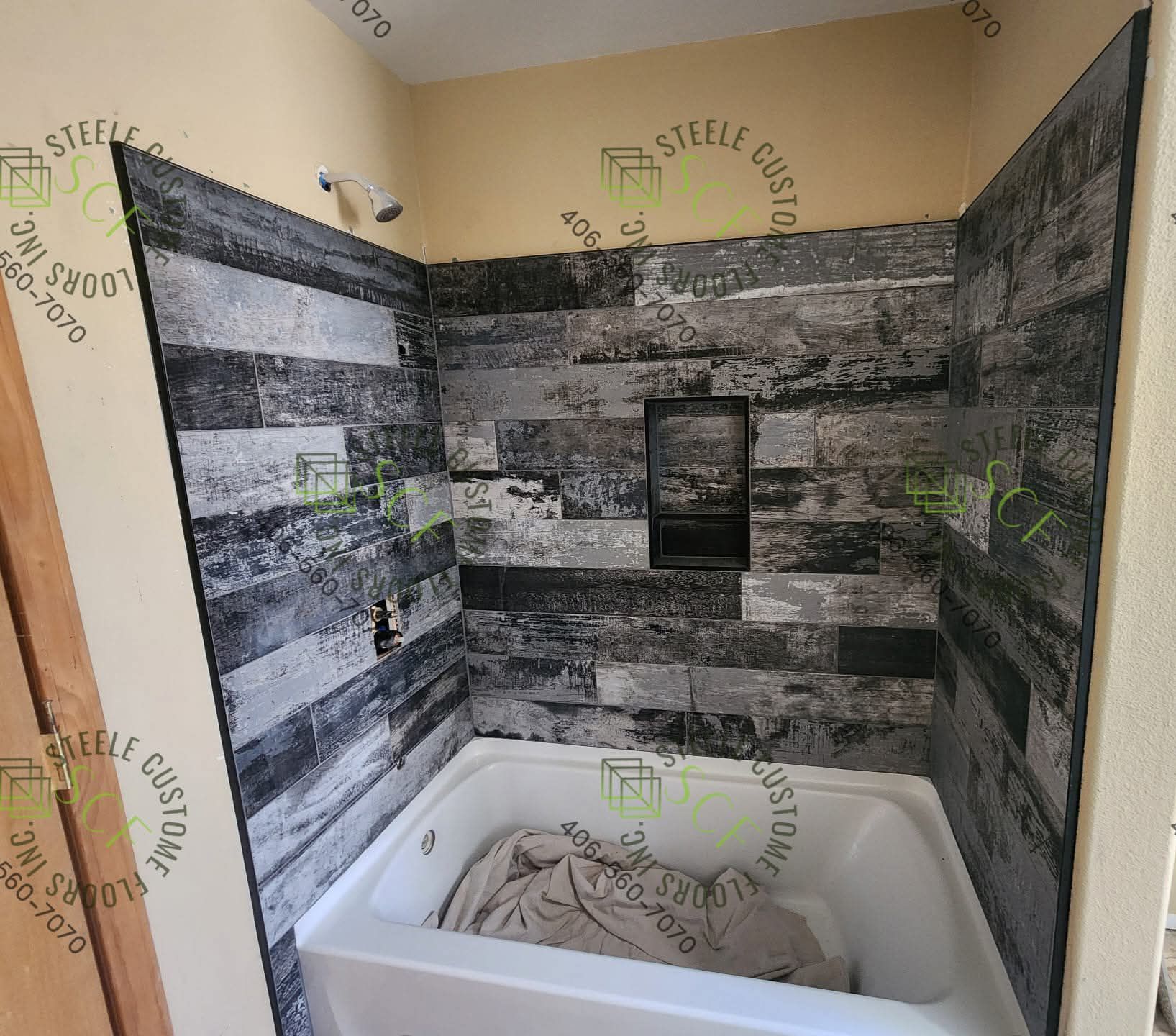 Shower stall with gray and black wood-look paneling, white tub, and a recessed shelf.