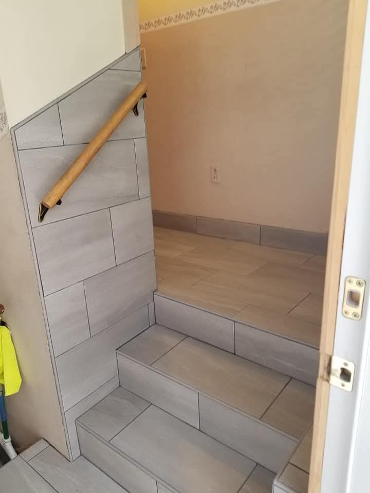 Staircase with light gray tile steps and wall, tan handrail, and doorway to a tan room.