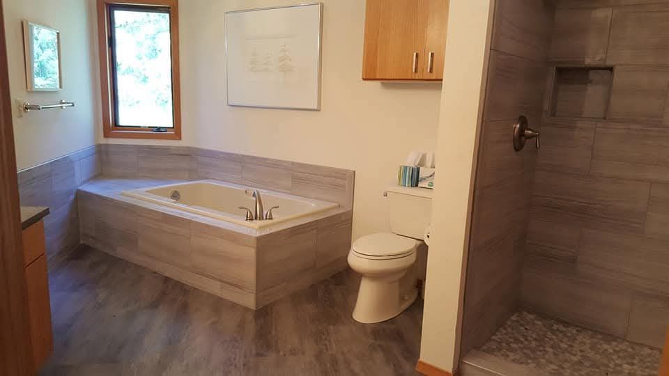 Bathroom with a built-in tub, tiled shower, toilet, window, and wood cabinetry. Gray and beige tones dominate.