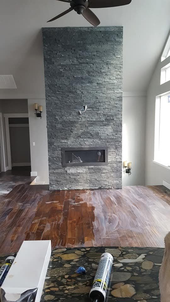Living room with stone fireplace, wood floor partially refinished, and ceiling fan.