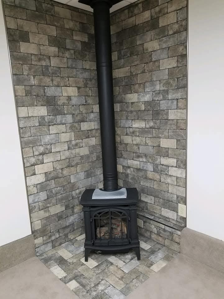 Black wood-burning stove in a corner with a brick-like wall and a black chimney.