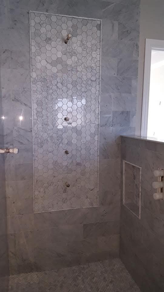 Gray tiled shower with a white hexagon tile accent wall featuring showerheads and a recessed shelf.