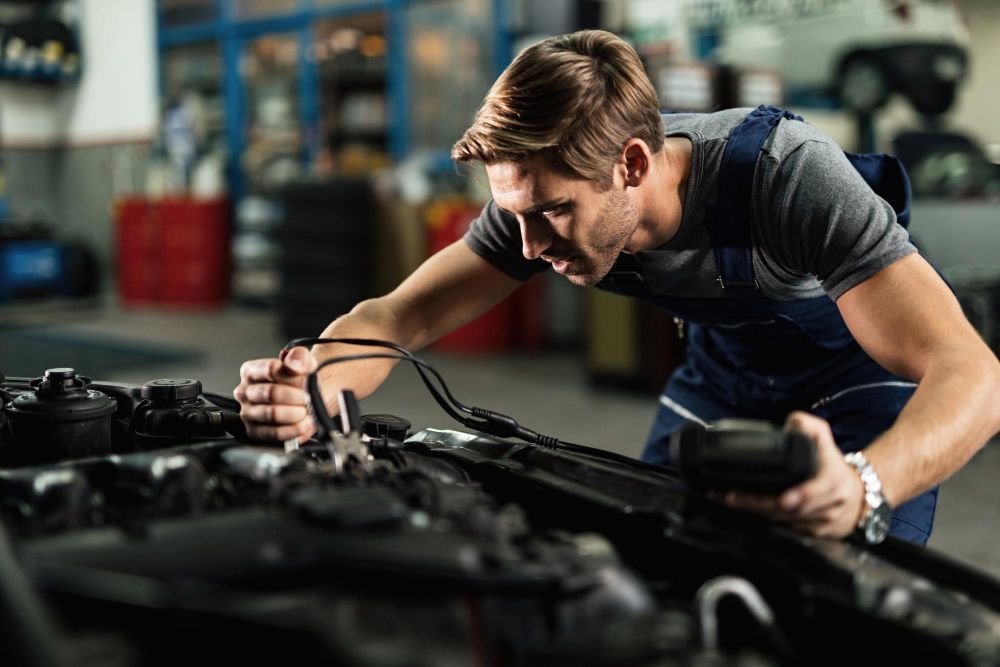 Mechanic inspecting a car engine in a workshop, leaning over the hood with tools.
