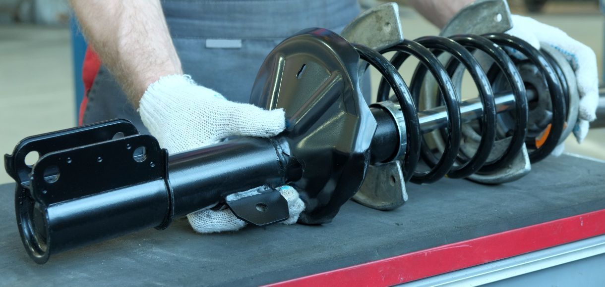 Mechanic's gloved hands handling a black suspension strut with coil spring on a workbench