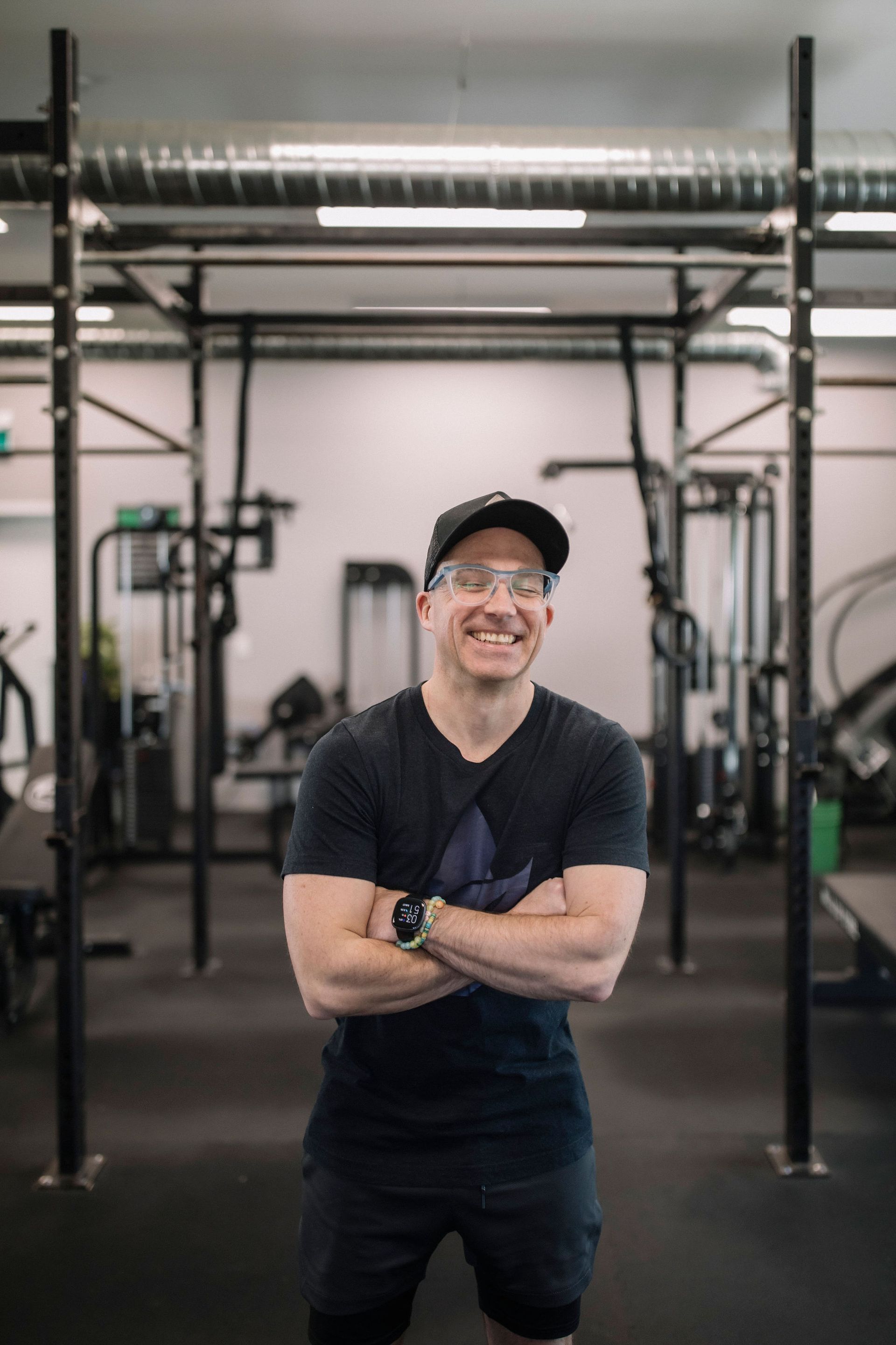 A man wearing a hat and glasses is standing with his arms crossed in a gym.