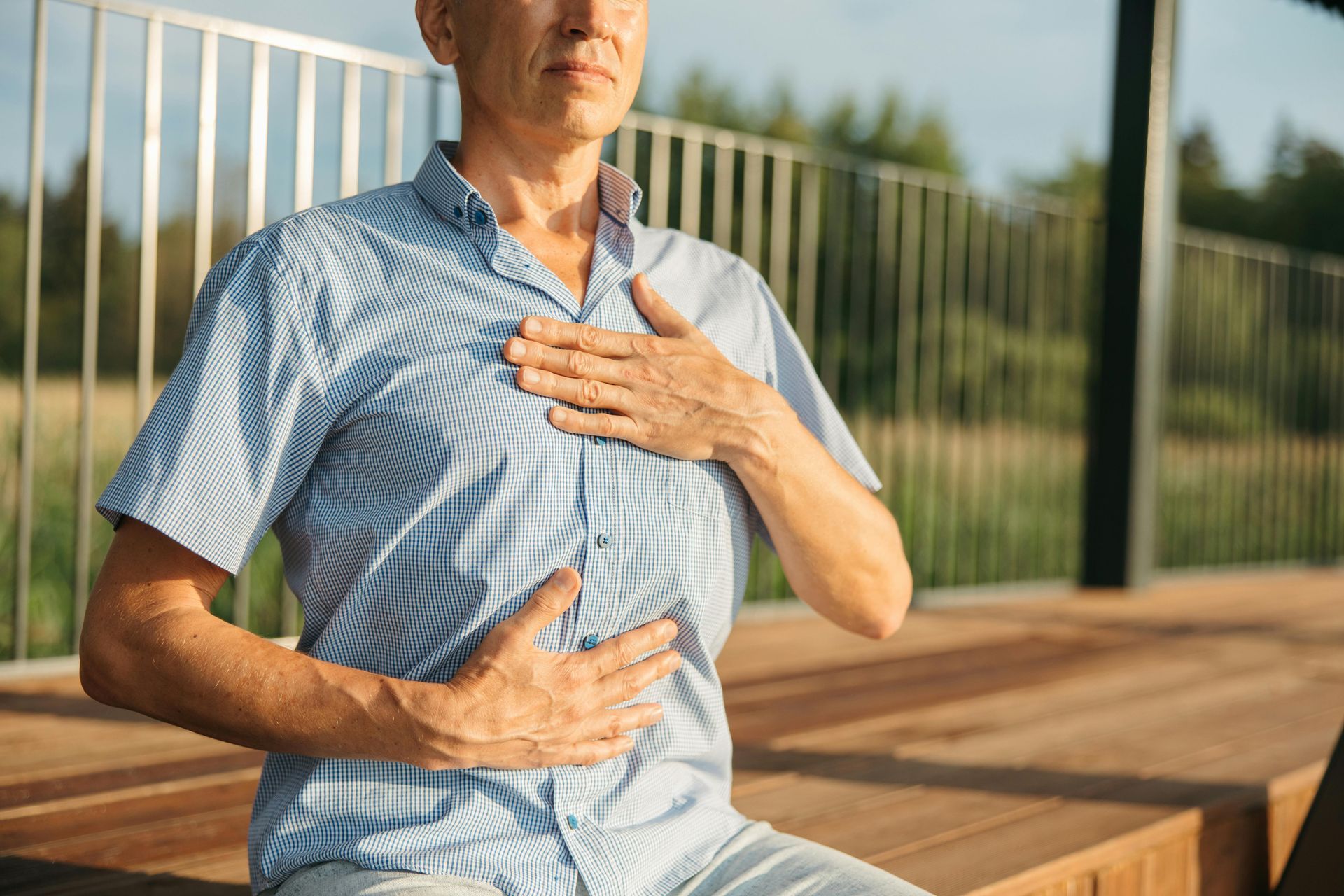Person seated outdoors holding their chest and abdomen, with a railing and wooden deck in the background.