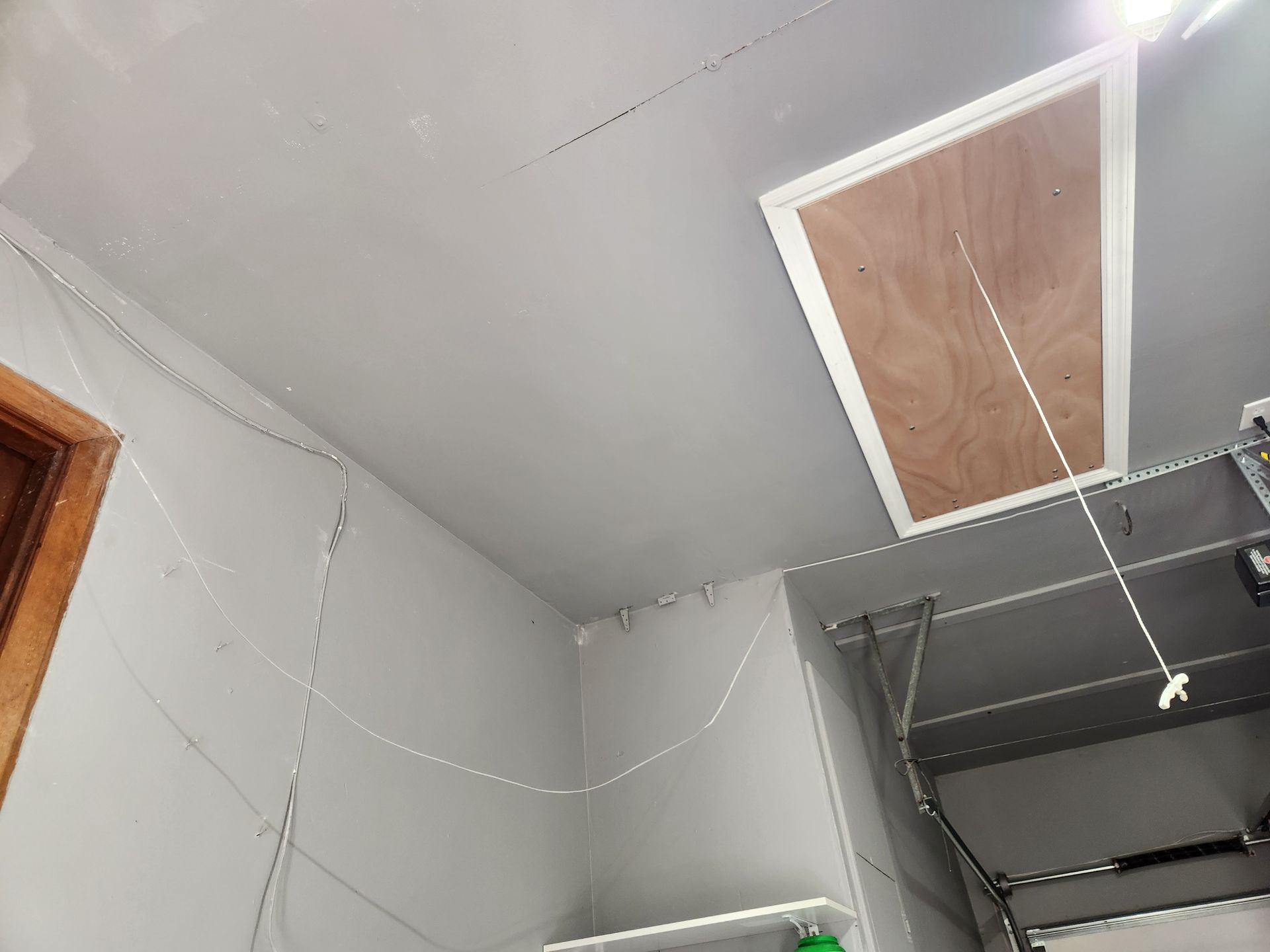 Garage interior with attic access door open. White and grey walls, exposed wiring, and a closed garage door.