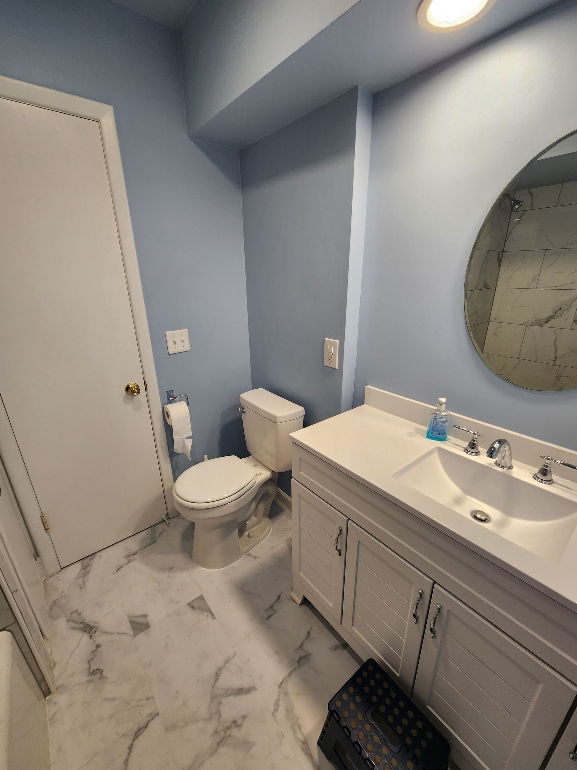 Bathroom with white vanity, toilet, round mirror, blue walls, and marble floor.