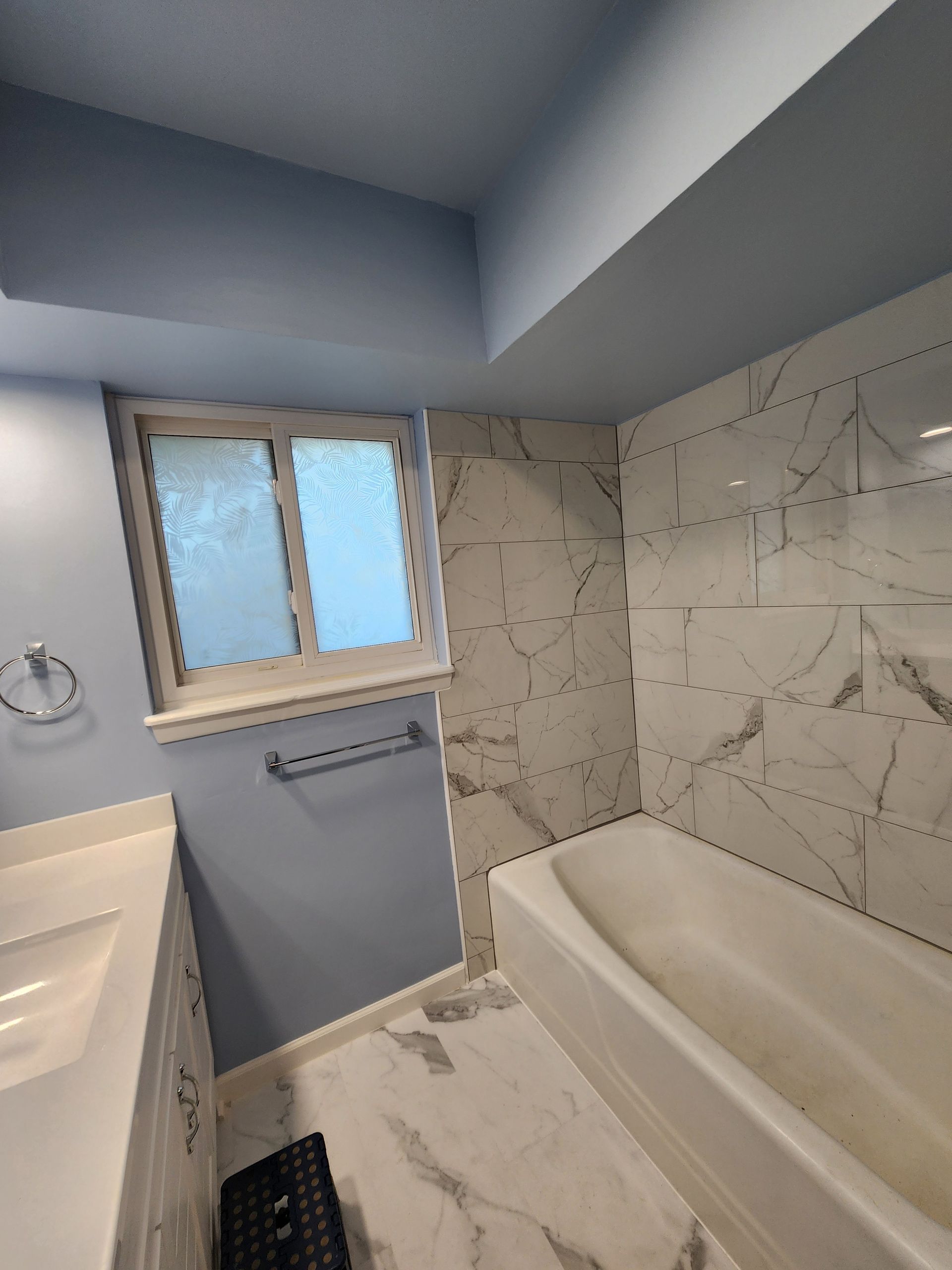 Bathroom with white marble-look tiles, blue walls, a bathtub, and a window with frosted glass.