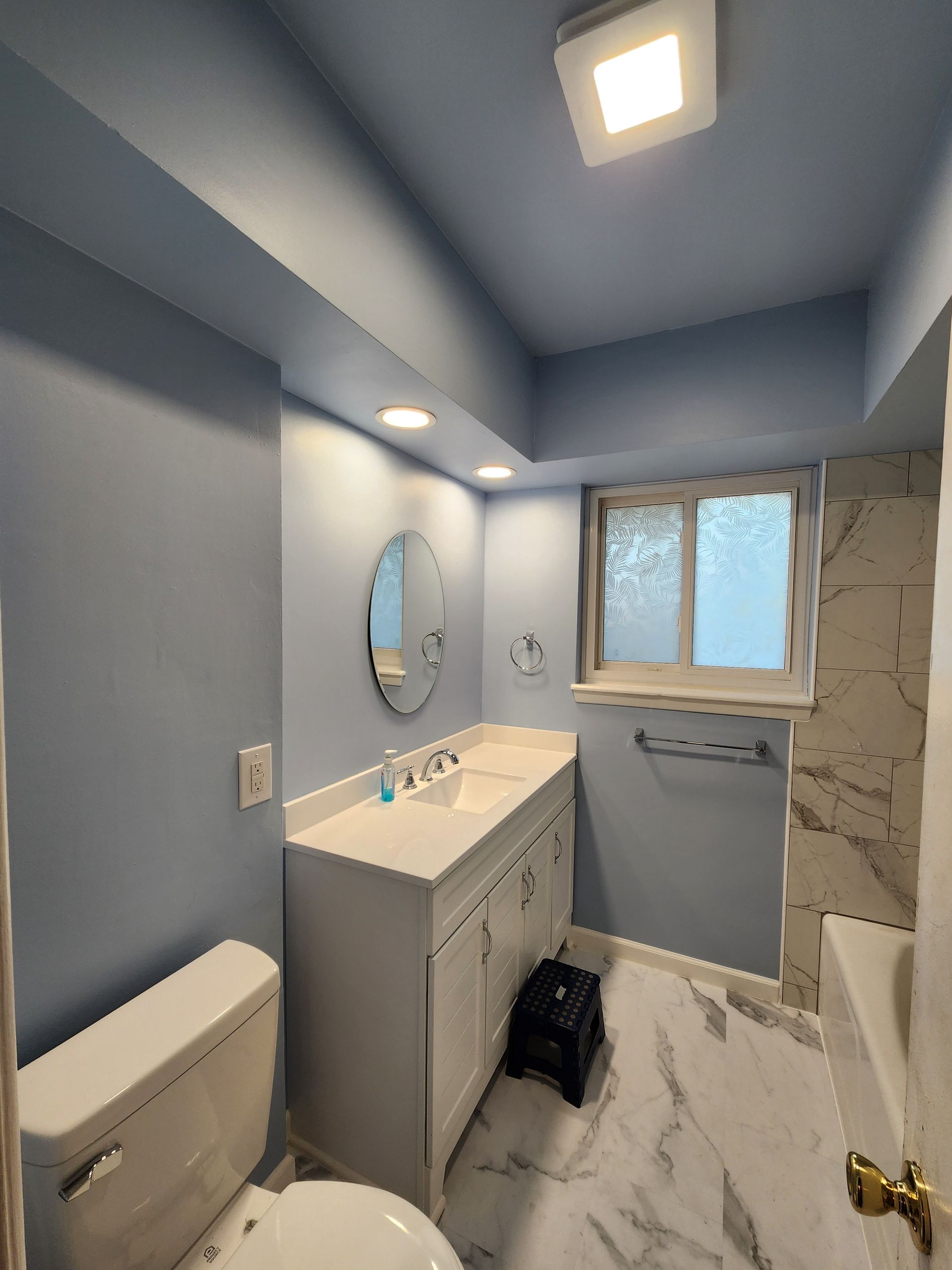 Blue bathroom with white vanity, toilet, and marble-look floor. Frosted window and recessed lighting.