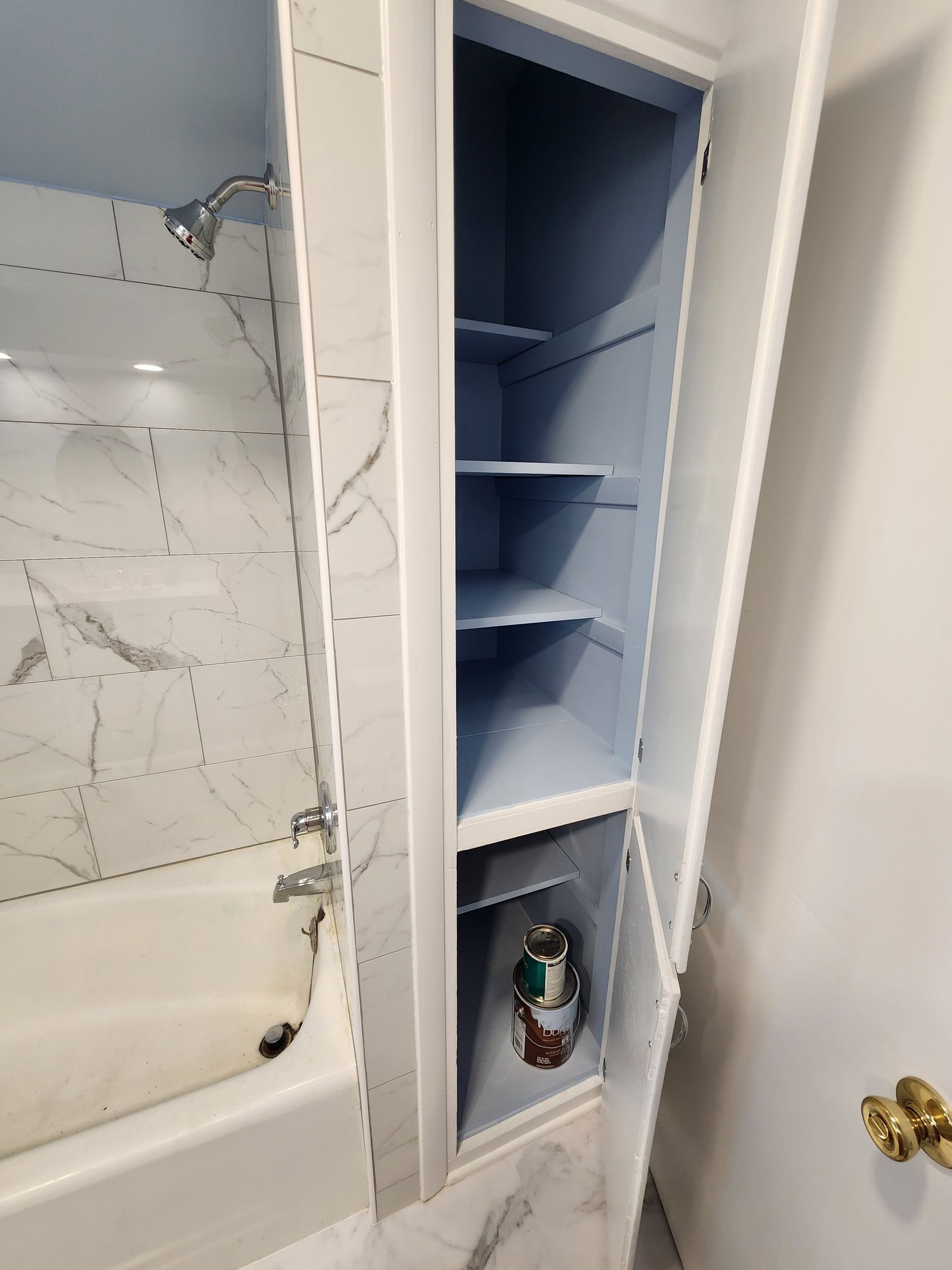 Bathroom cabinet with open door revealing shelves, paint can, and tub/shower. Blue interior, white exterior.