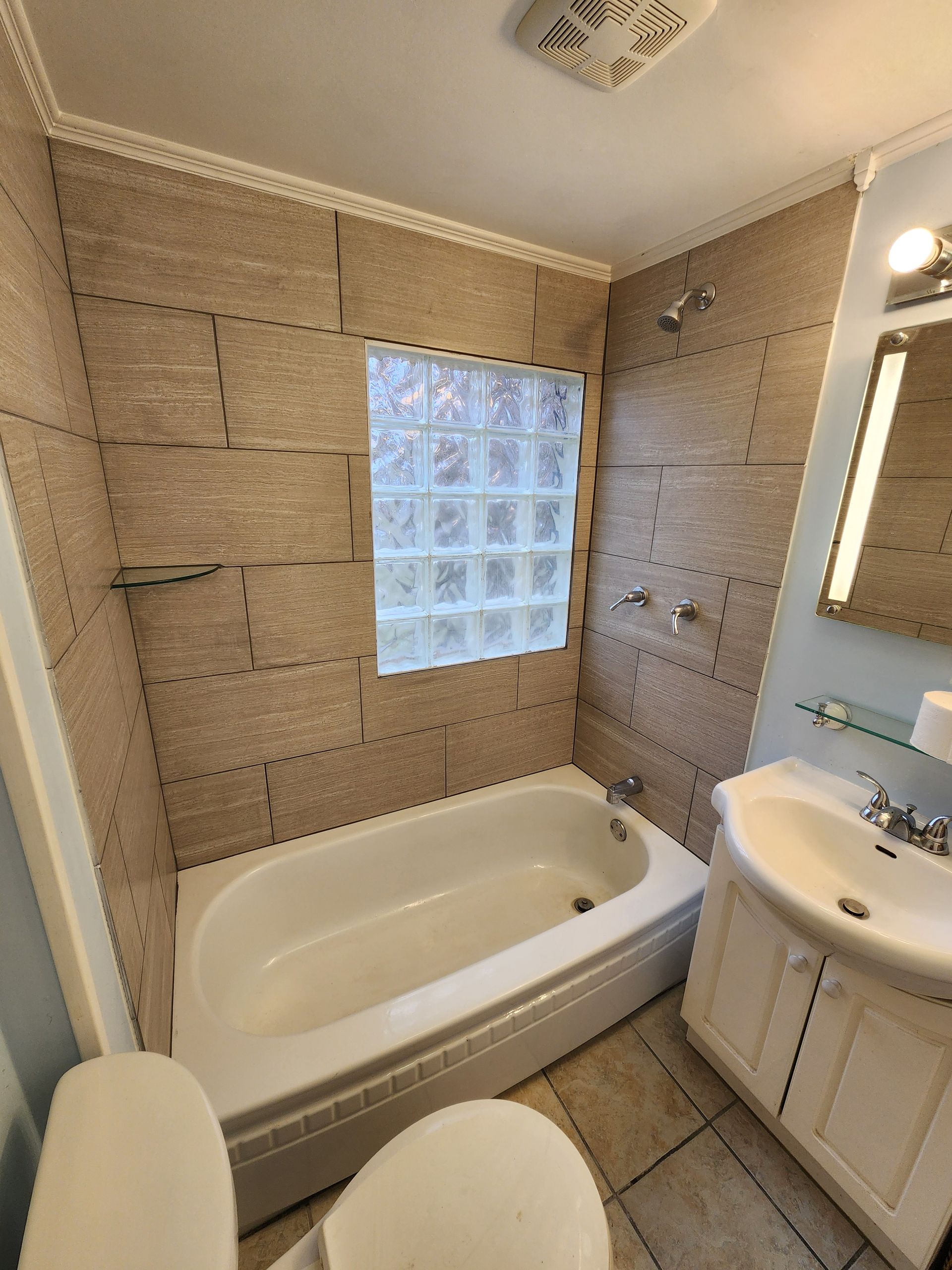 Bathroom with beige tile walls, white tub, toilet, and vanity, and a window with glass blocks.