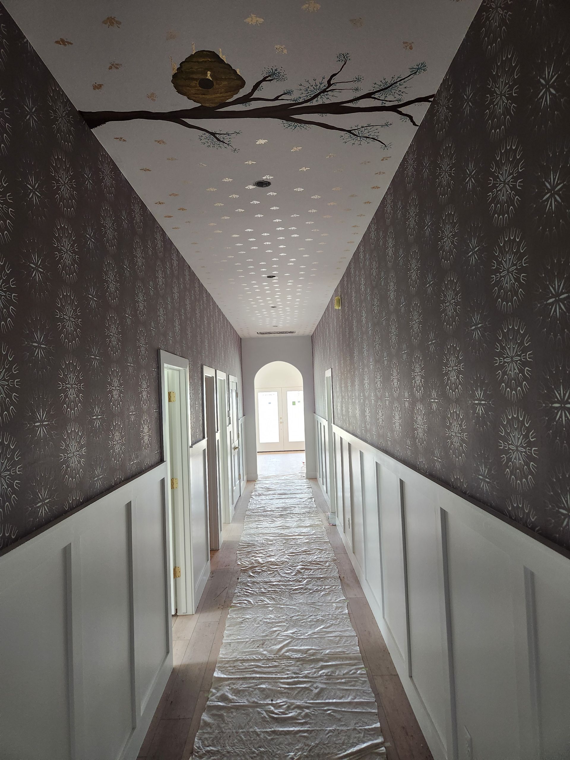 Long hallway with patterned wallpaper, white wainscoting, and a painted branch on the ceiling.