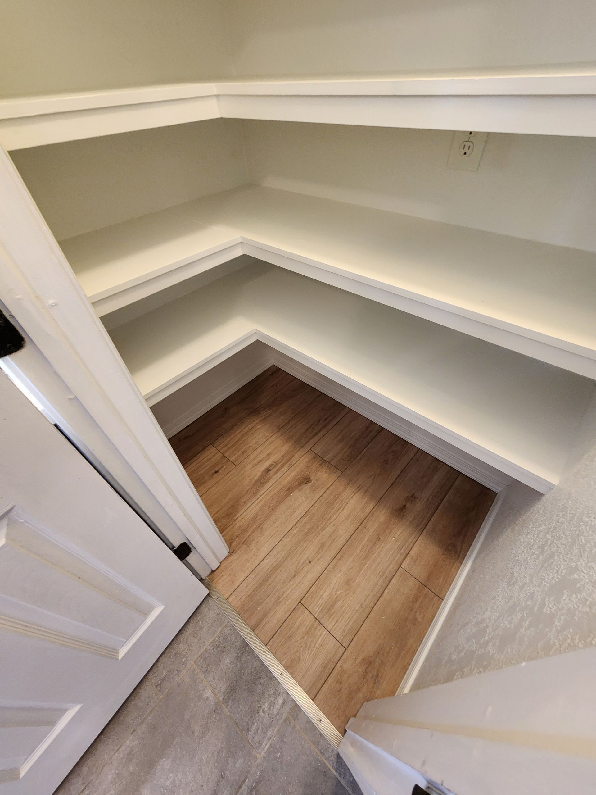 Empty corner pantry with built-in white shelves and wood-look flooring. Ajar white door.