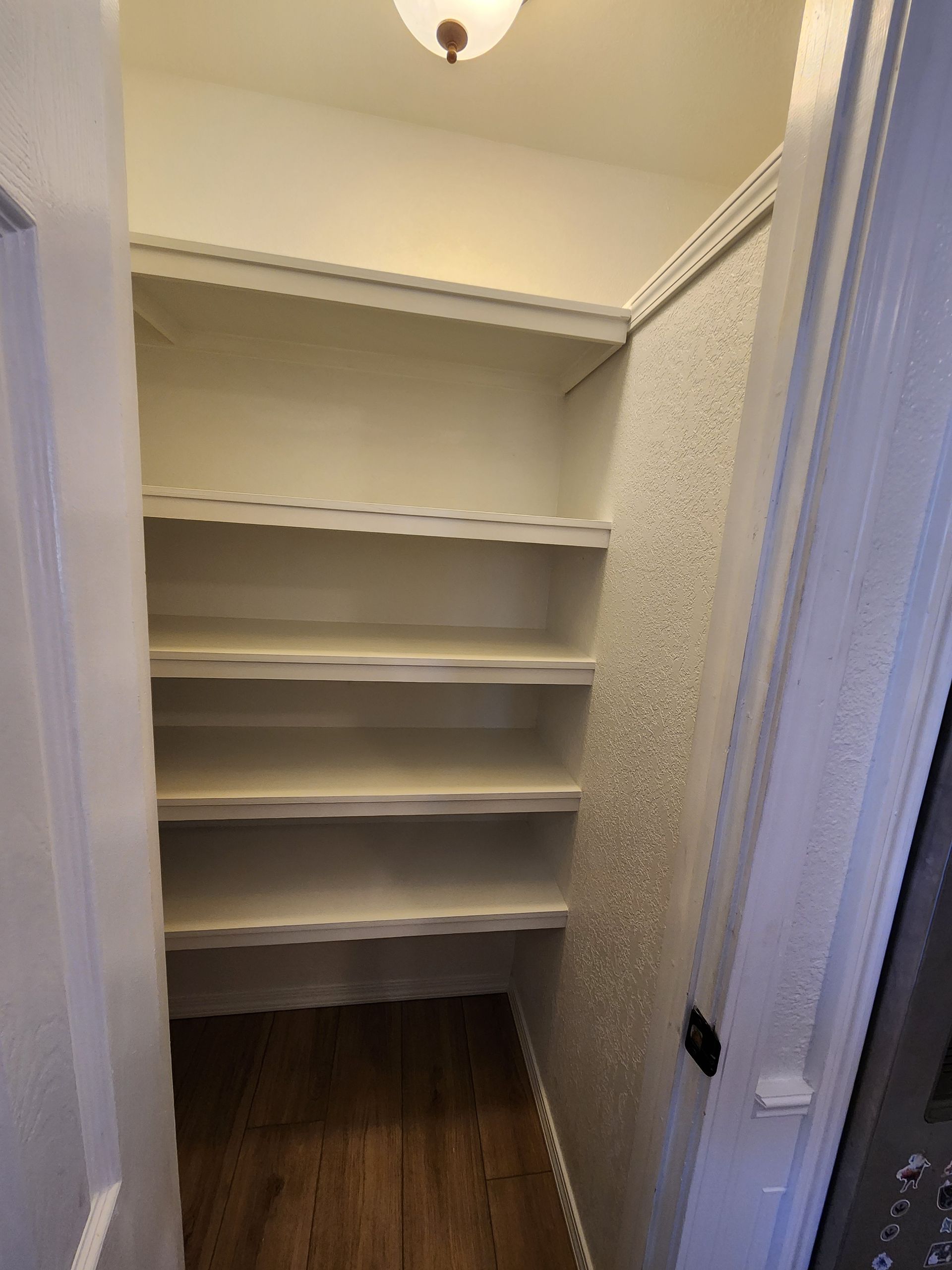 White pantry shelves in a narrow closet with wood flooring and a single overhead light.